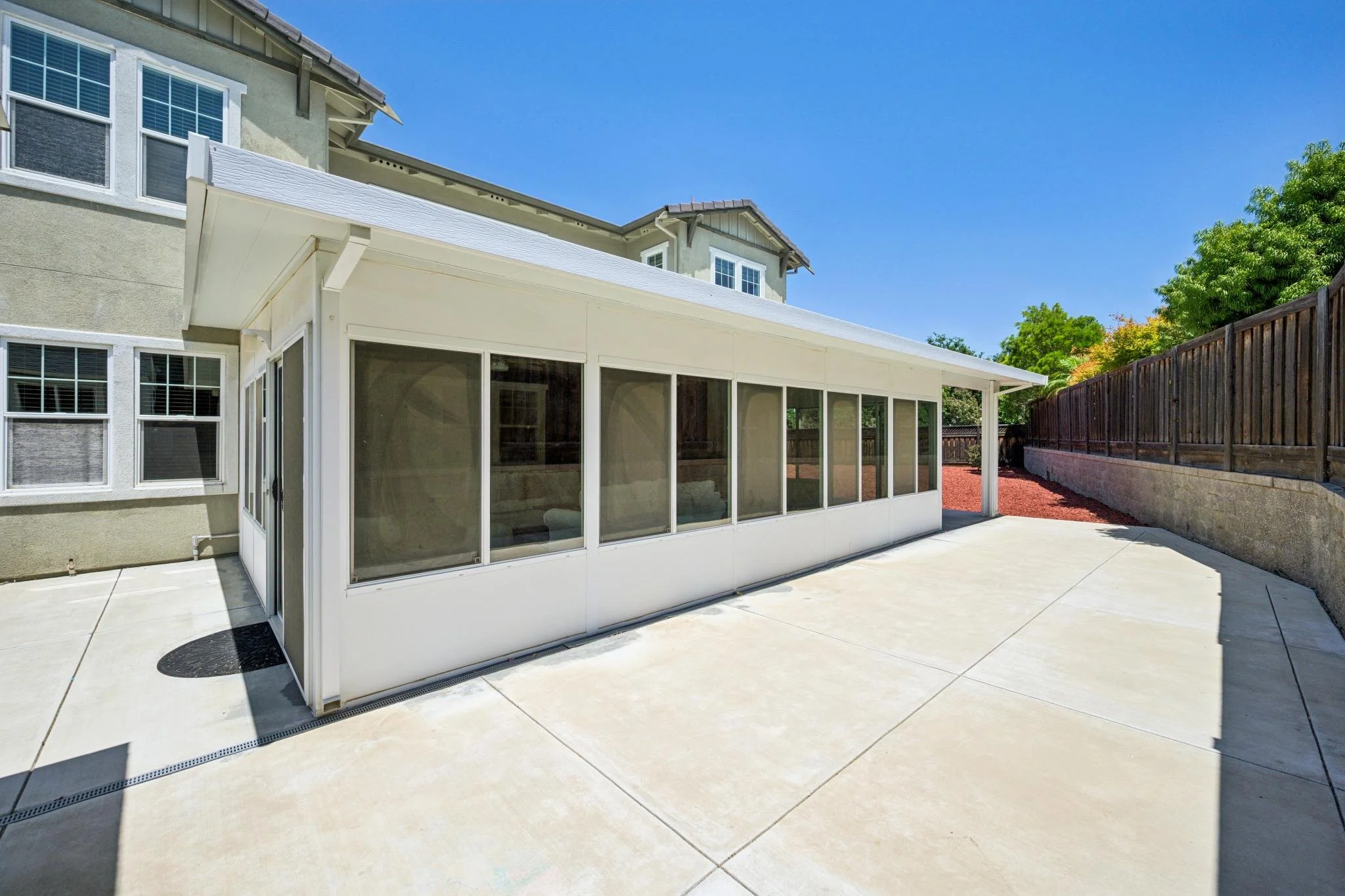 A backyard patio with a screened-in porch, concrete flooring, a wooden fence on one side, and a light green house with large windows and a gable roof in the background, under a clear blue sky.