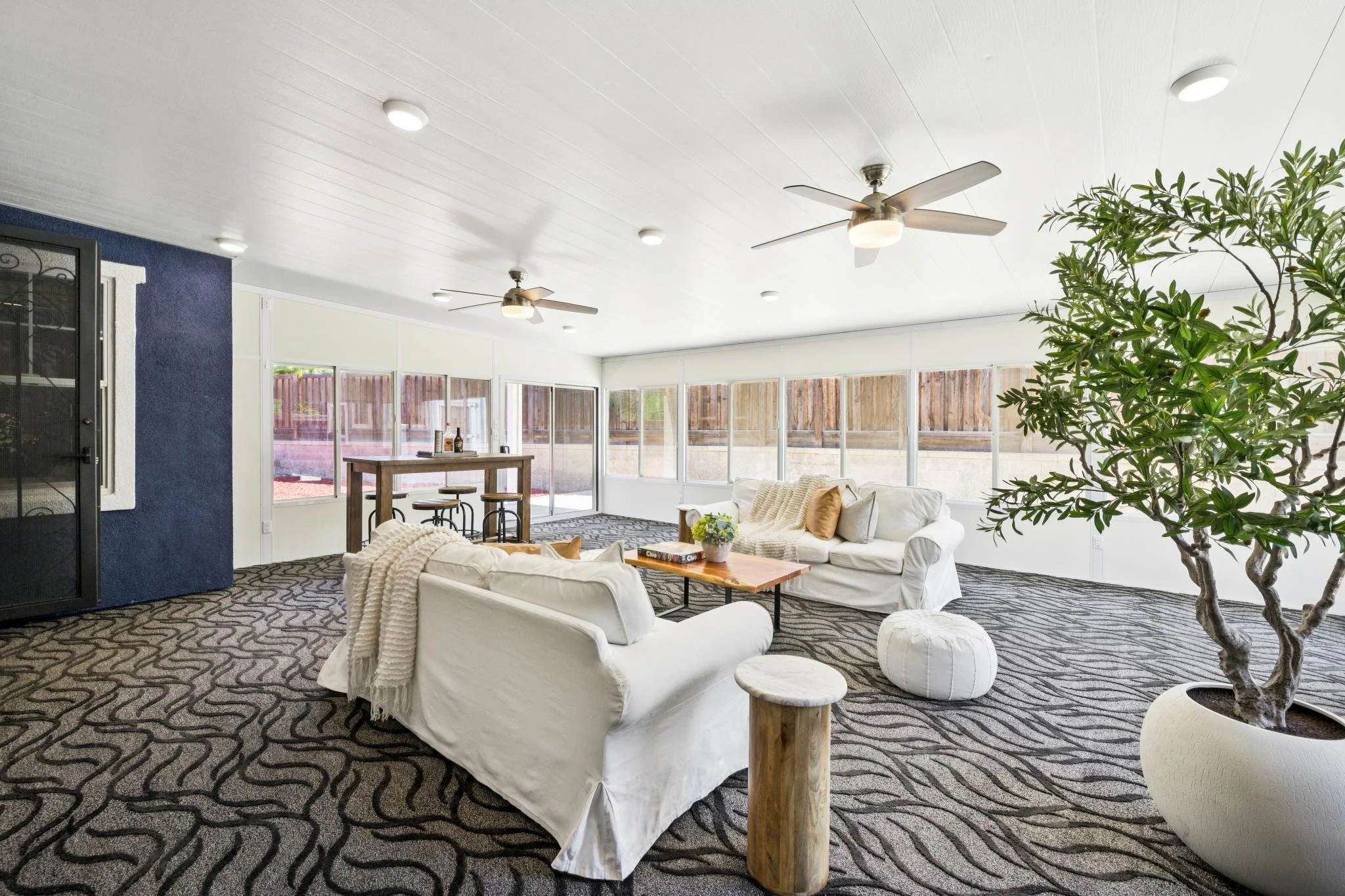 Bright living room with white sofas, patterned carpet, large windows, a ceiling fan, and a potted tree.