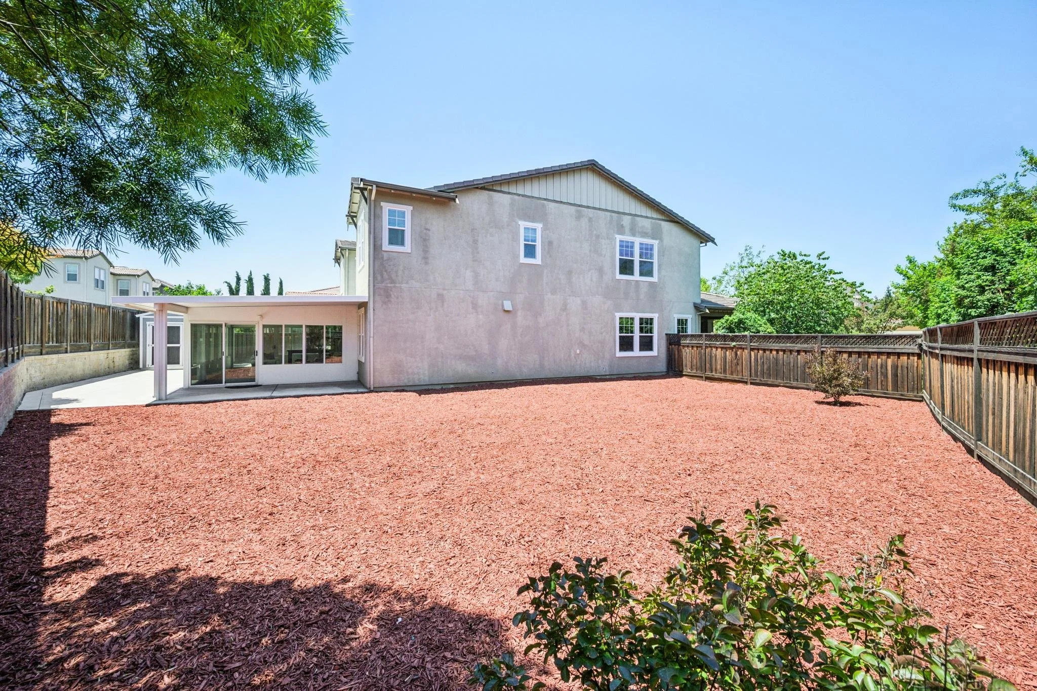 Backyard with reddish mulch, a small tree, and a two-story house with a patio and wood fence
