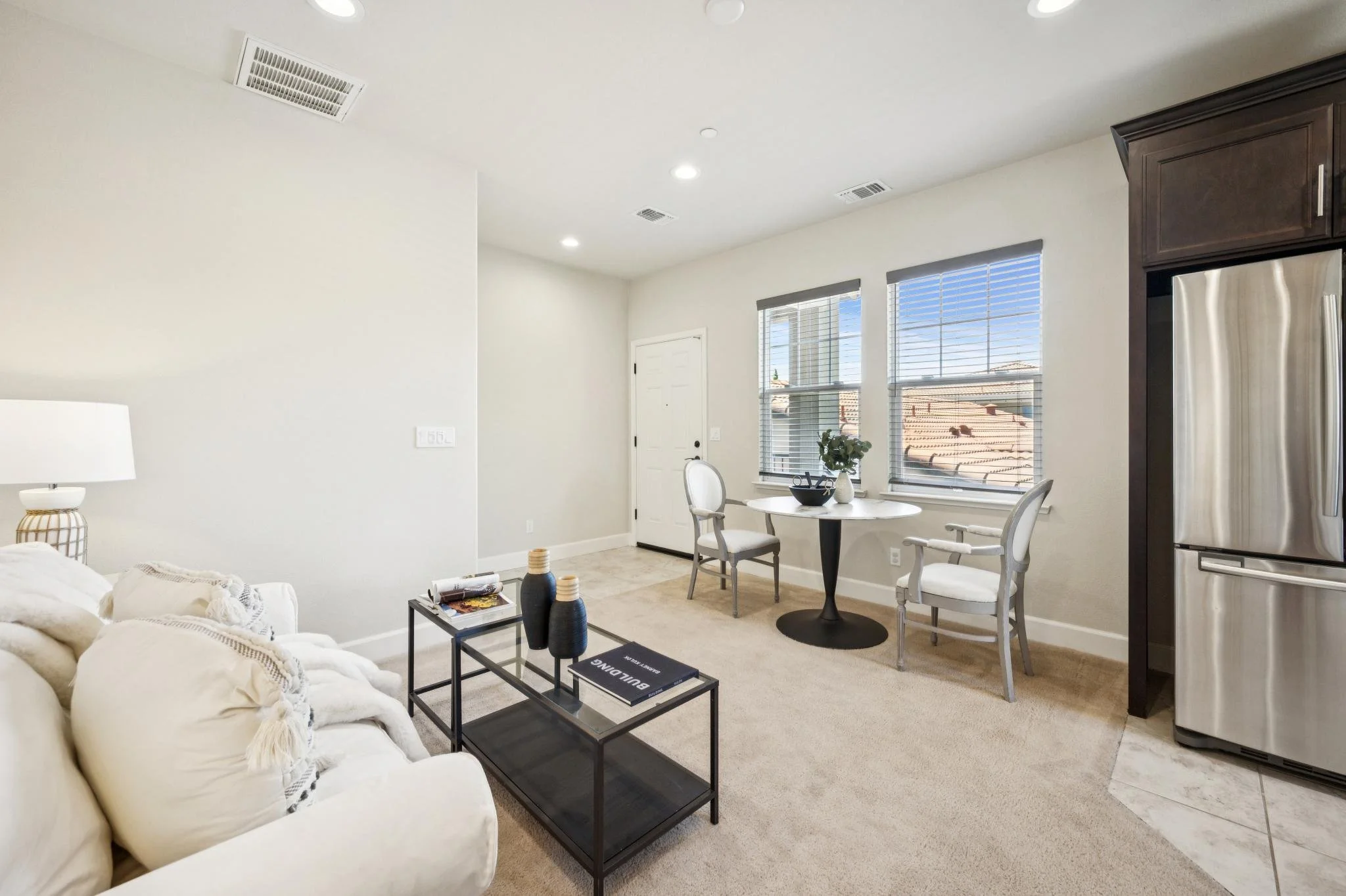 A bright living room with white walls and beige carpet. There is a white sofa with neutral pillows, a black coffee table with decorative vases and magazines, a small white round dining table with two chairs near large windows, and a dark wood refrige