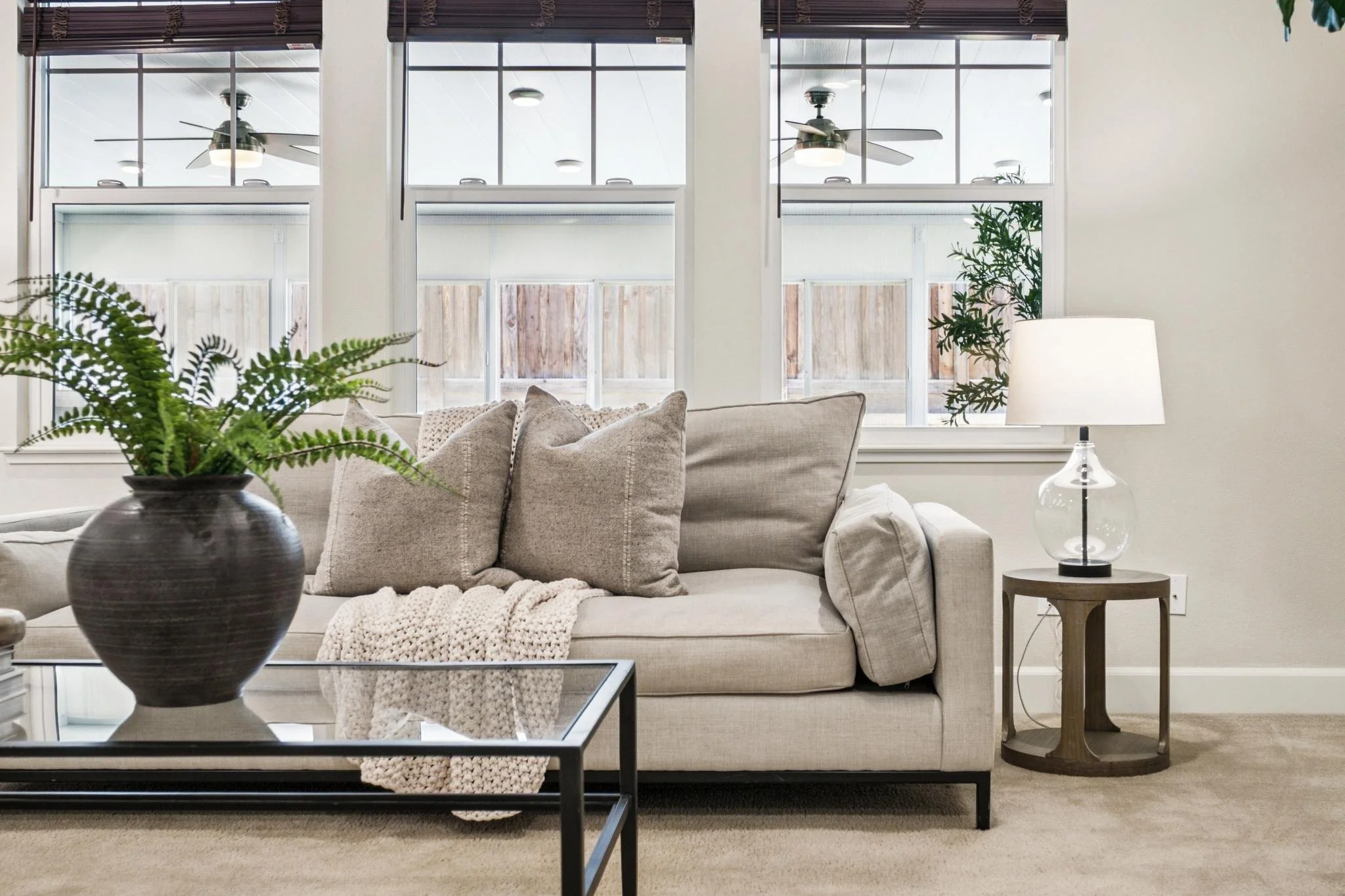 Living room with beige sofa, decorative pillows, a blanket, a round wooden side table with a lamp, a large potted plant, and a coffee table with a glass top. Behind the sofa, three large windows with wooden blinds are visible, with ceiling fans outsi