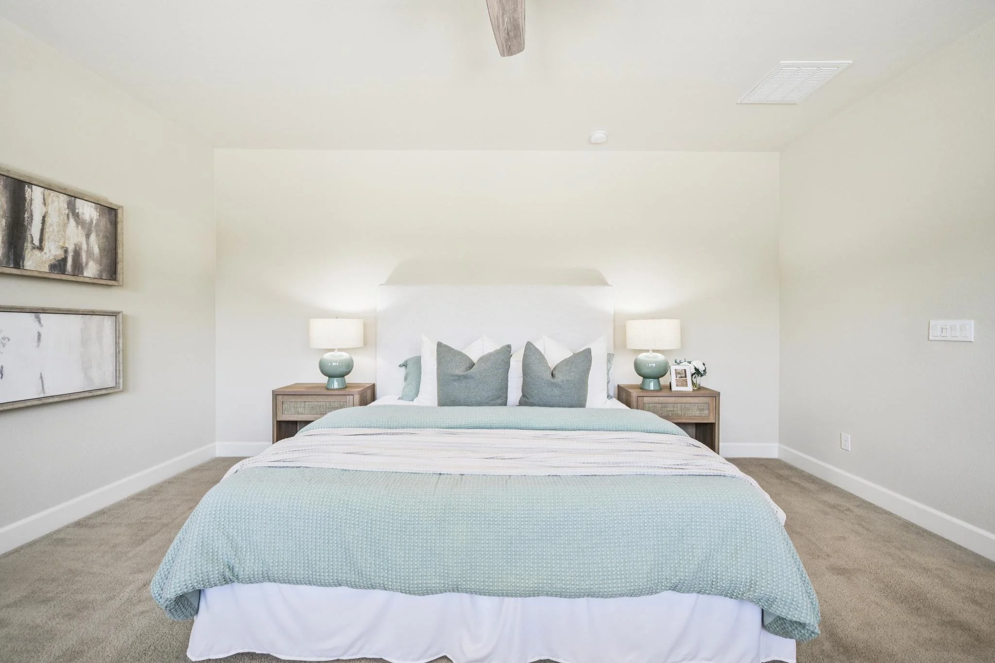 A neatly made bed with a light blue textured bedspread, decorated with multiple pillows, in a white-walled bedroom. Two wooden nightstands with white lamps and small decorative items flank the bed. Wall art hangs on the left side of the room.