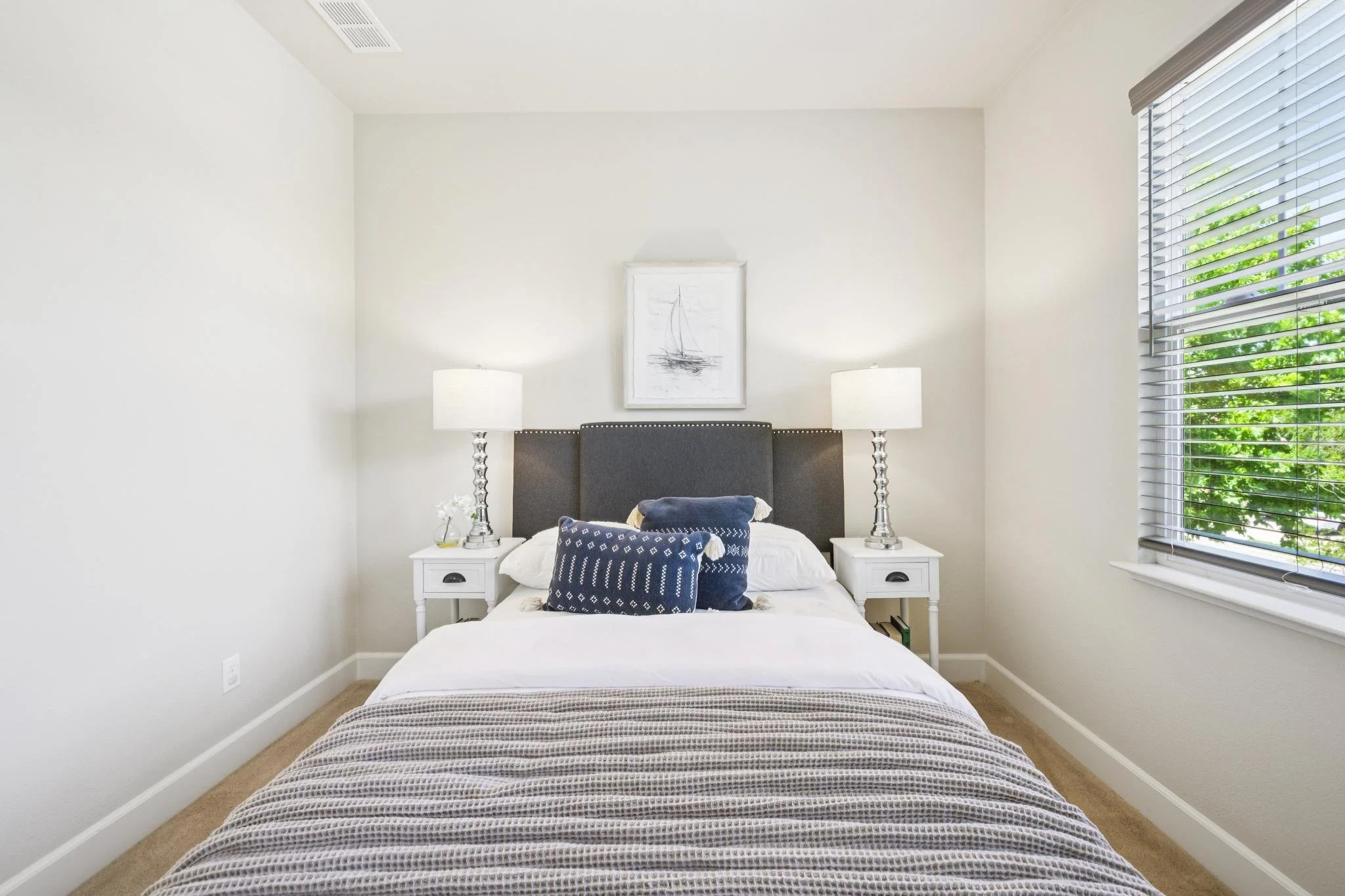 A bedroom with a white bed, two white nightstands with lamps, a navy blue and white decorative pillow, a framed sailboat painting above the dark gray headboard, a window with blinds showing green trees outside, and beige carpeted floor.