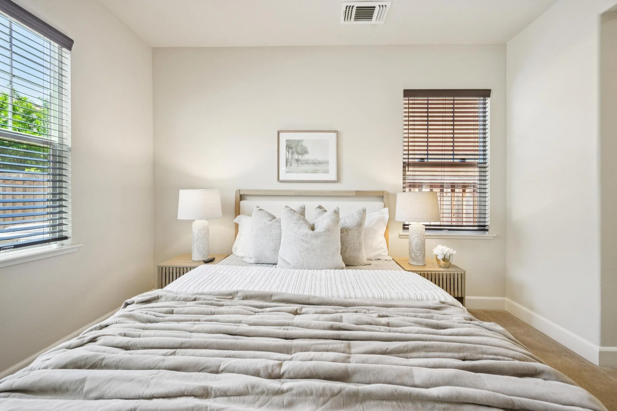 A bedroom with two windows, a bed with pillows, two lamps on bedside tables, a framed picture on the wall, a small plant, and light-colored walls.