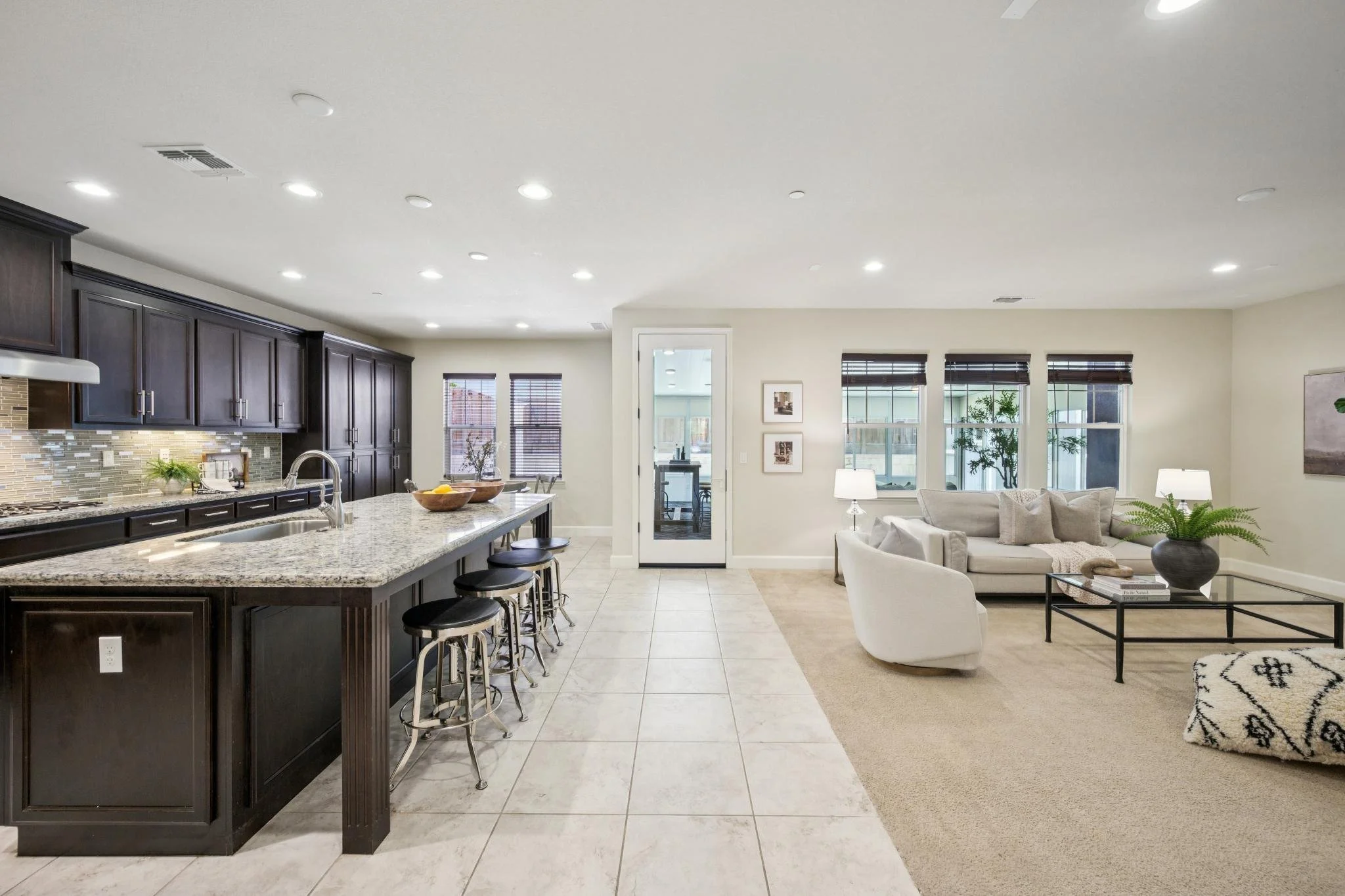 Open-concept kitchen and living room with dark wood cabinets, granite countertops, bar stools, beige tiled floor, and a seating area with white chairs and sofa, windows with blinds, and potted plants.