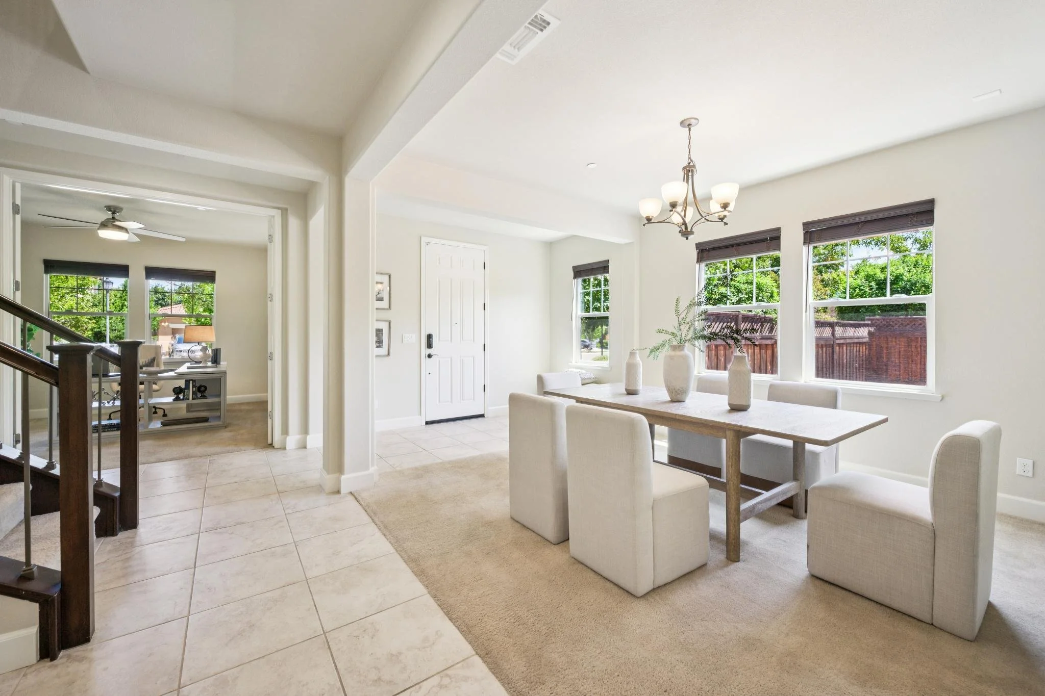 Inside view of a bright living and dining area with large windows showing green trees outside, white walls, a chandelier, and a white dining table with chairs and decorative vases.