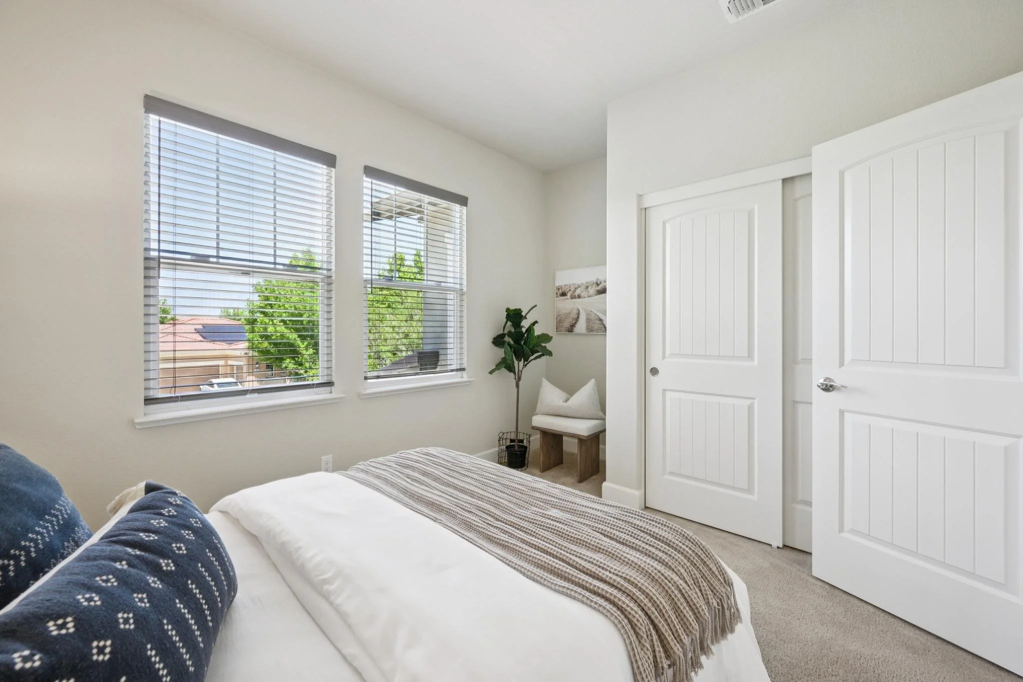 A bedroom with a white bed, dark blue patterned pillows, a striped blanket, a small padded bench beside the bed, and large windows with blinds showing trees and a neighboring house outside.