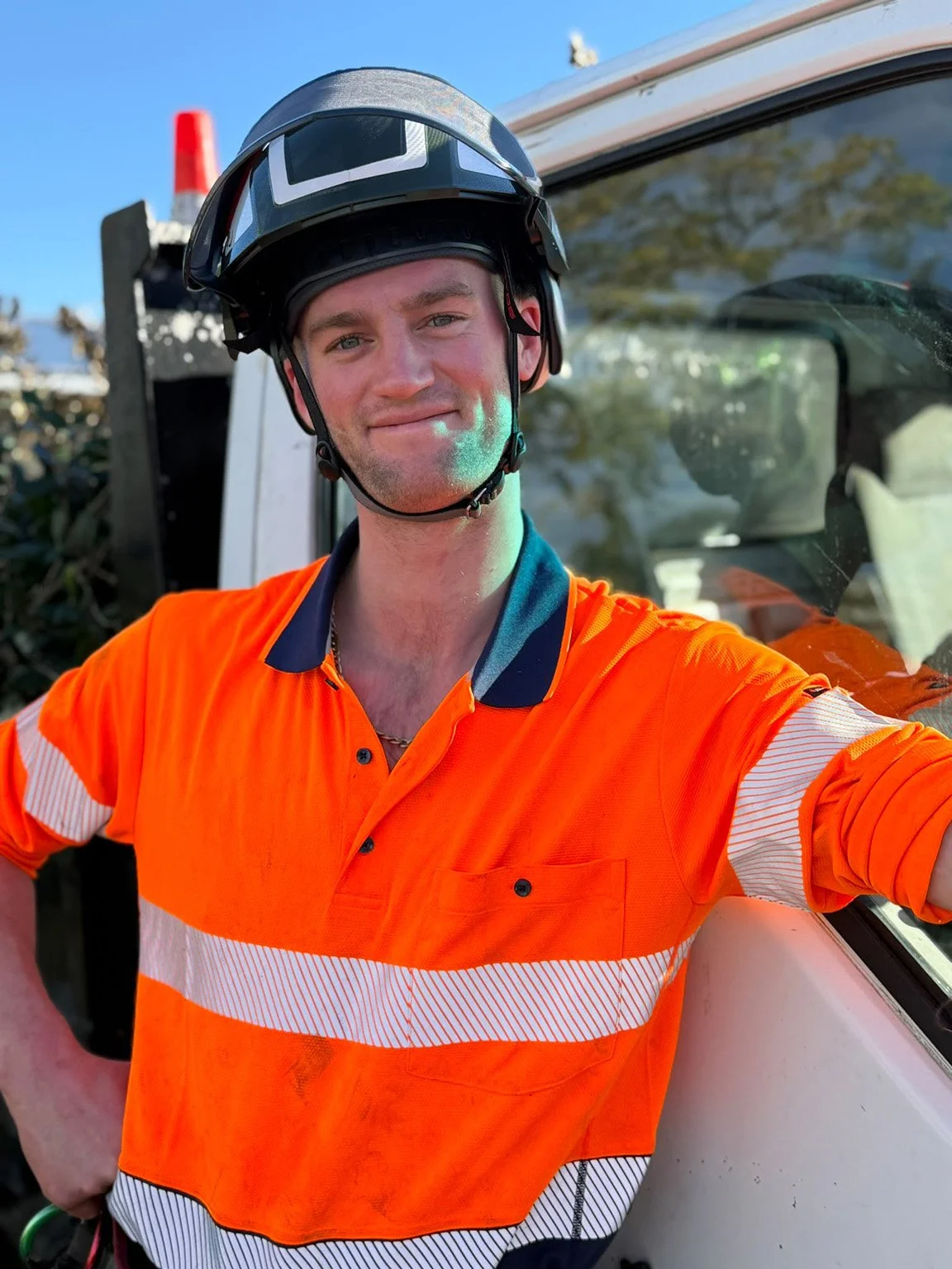 A man wearing a black safety helmet and orange high-visibility safety shirt takes a selfie outdoors during the daytime, with a vehicle and trees visible in the background.