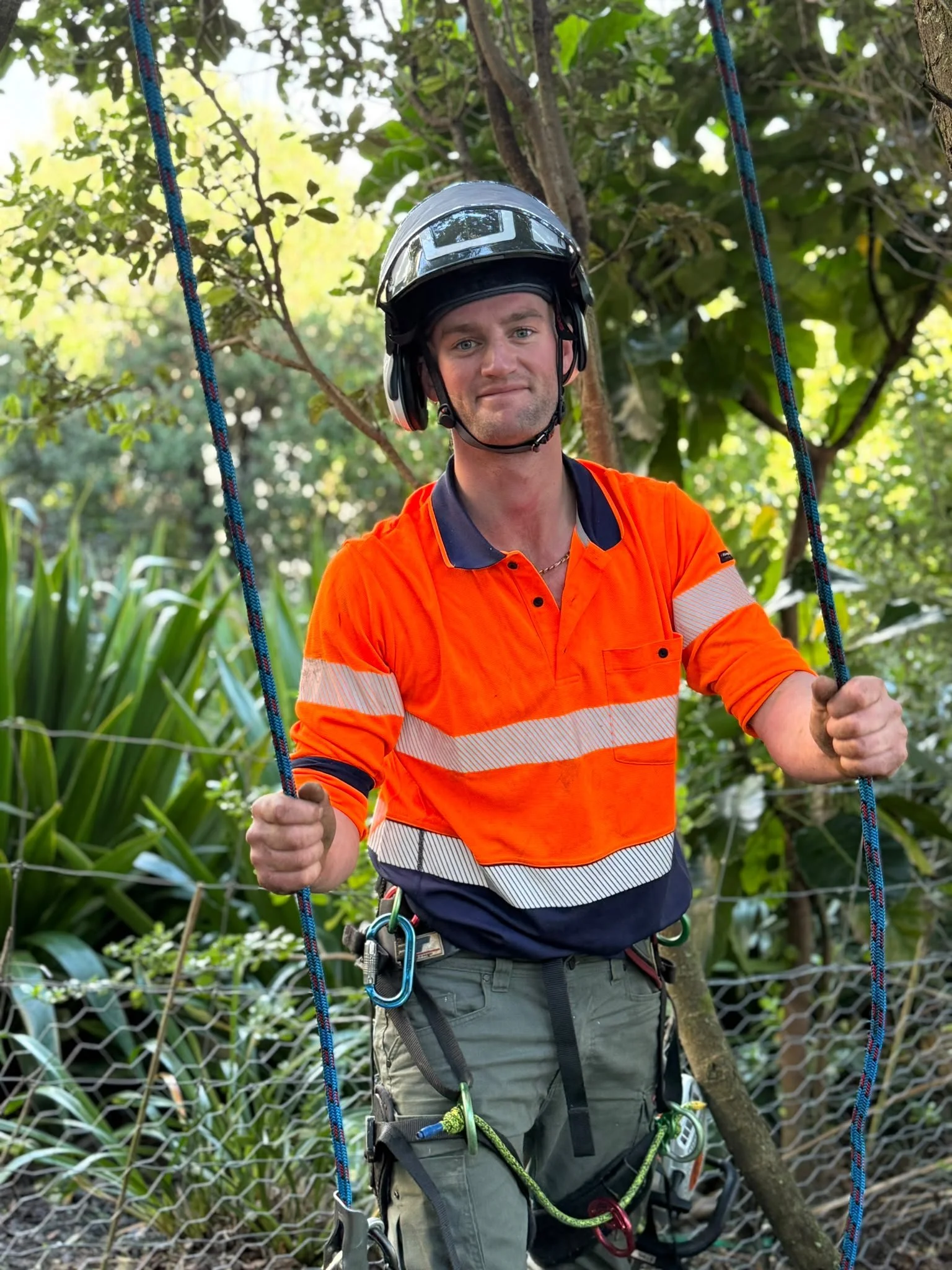Man wearing a helmet and orange safety shirt standing outdoors on a rope bridge, holding onto the ropes, surrounded by green foliage.