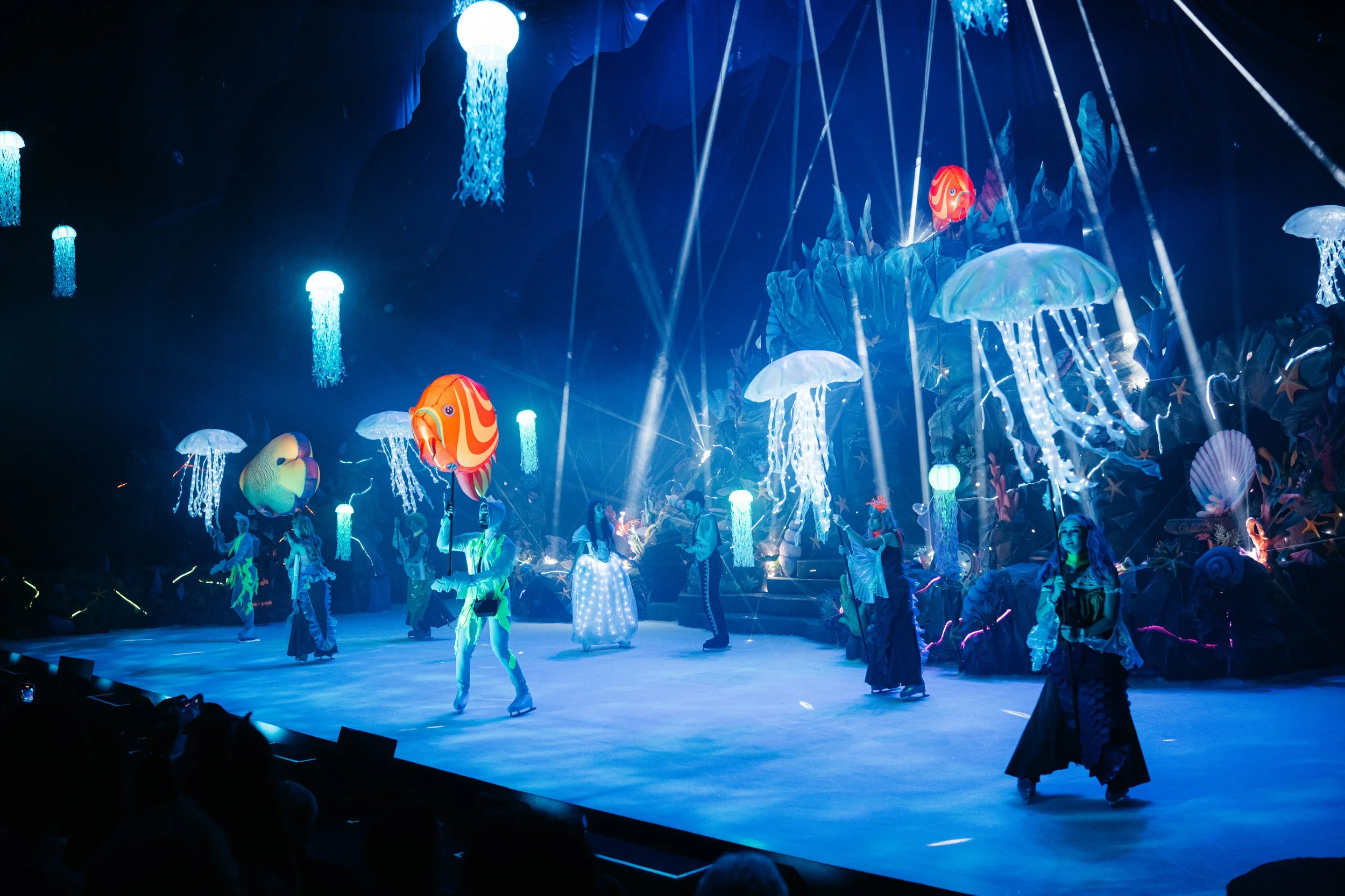 Cena de "A Pequena Sereia No Gelo" com artistas em palco rodeados por medusas iluminadas, peixes coloridos e elementos inspirados no fundo do mar. A iluminação em tons de azul cria uma atmosfera imersiva que transporta o público para o universo mágic