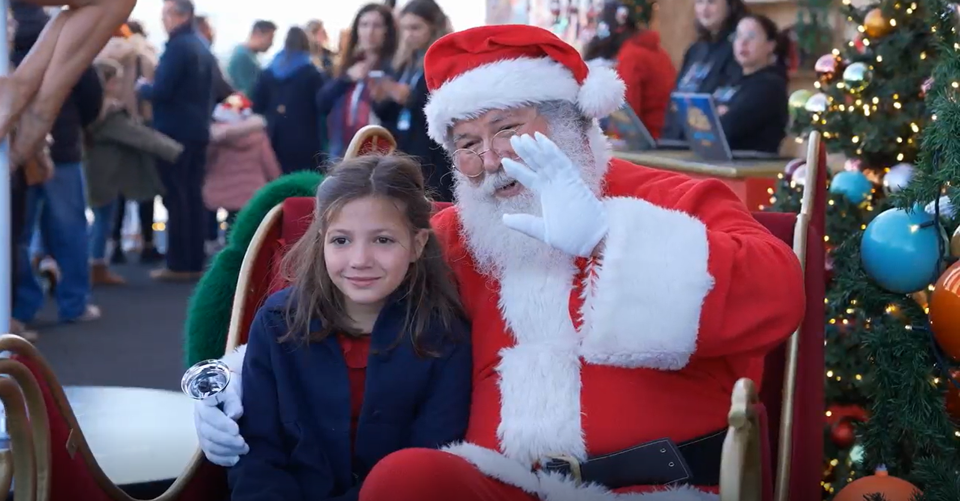 Uma menina de cabelo castanho claro e roupa azul sentada no colo do Papai Noel, que está vestindo de vermelho e branco, em um evento natalino. Ao lado deles, uma árvore de Natal decorada com enfeites coloridos e luzes.