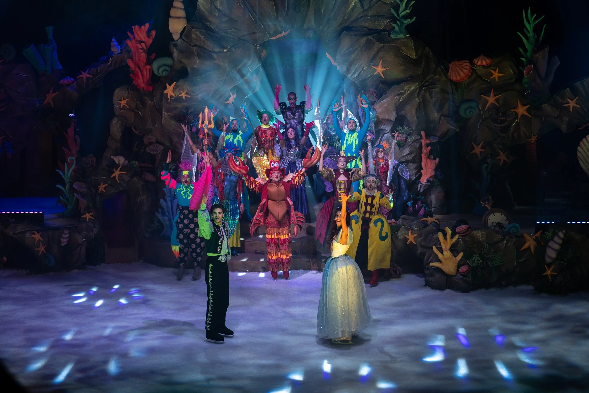 Momento de "A Pequena Sereia No Gelo" com Ariel e o Príncipe Eduard no centro da ação, enquanto as restantes personagens se reúnem num cenário submarino colorido, com corais, estrelas-do-mar e iluminação teatral.