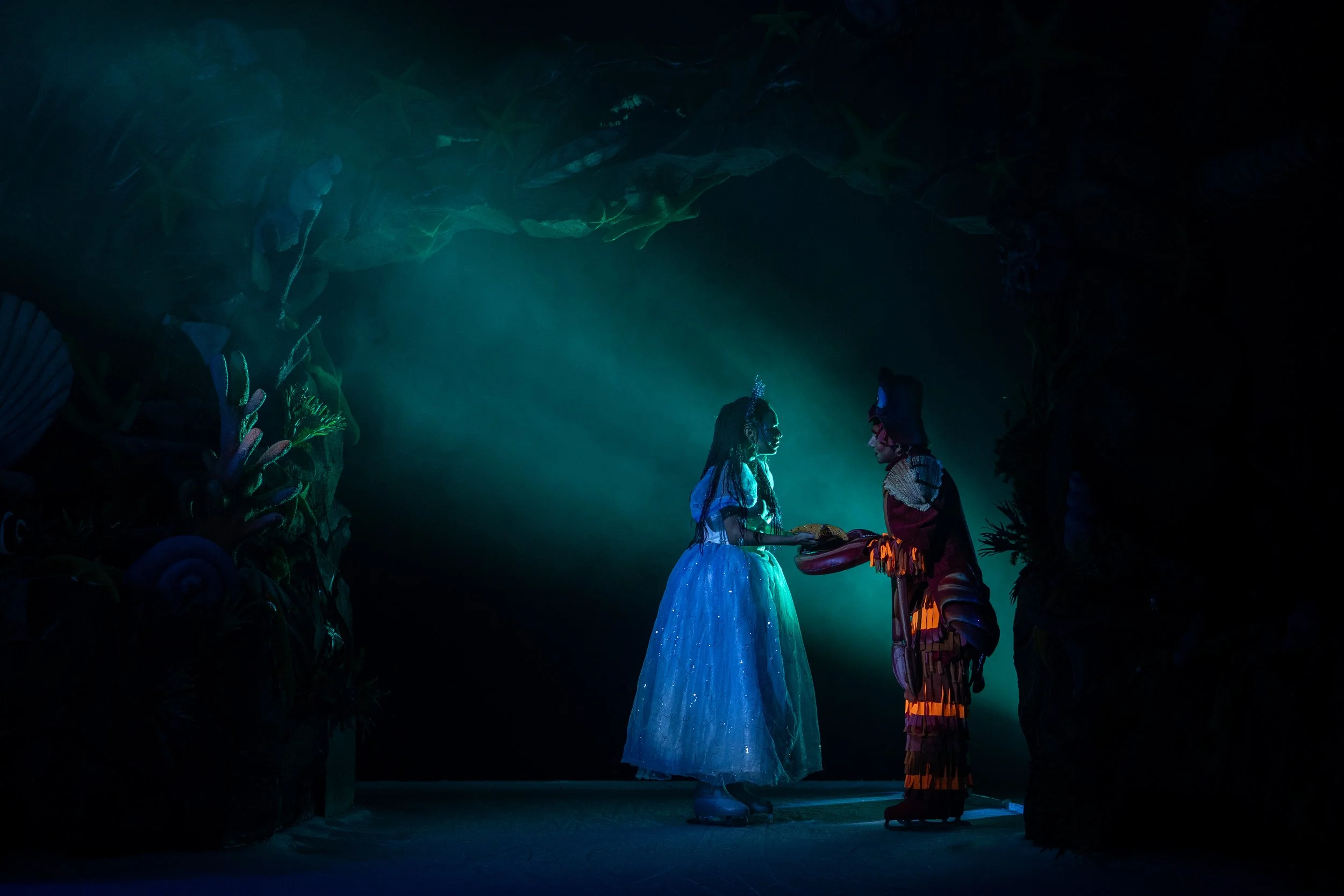 Cena de "A Pequena Sereia No Gelo" com Ariel e Horácio Crustáceo em palco, num momento de interação sob uma iluminação cénica em tons de azul e verde que recria a atmosfera mágica do fundo do mar.