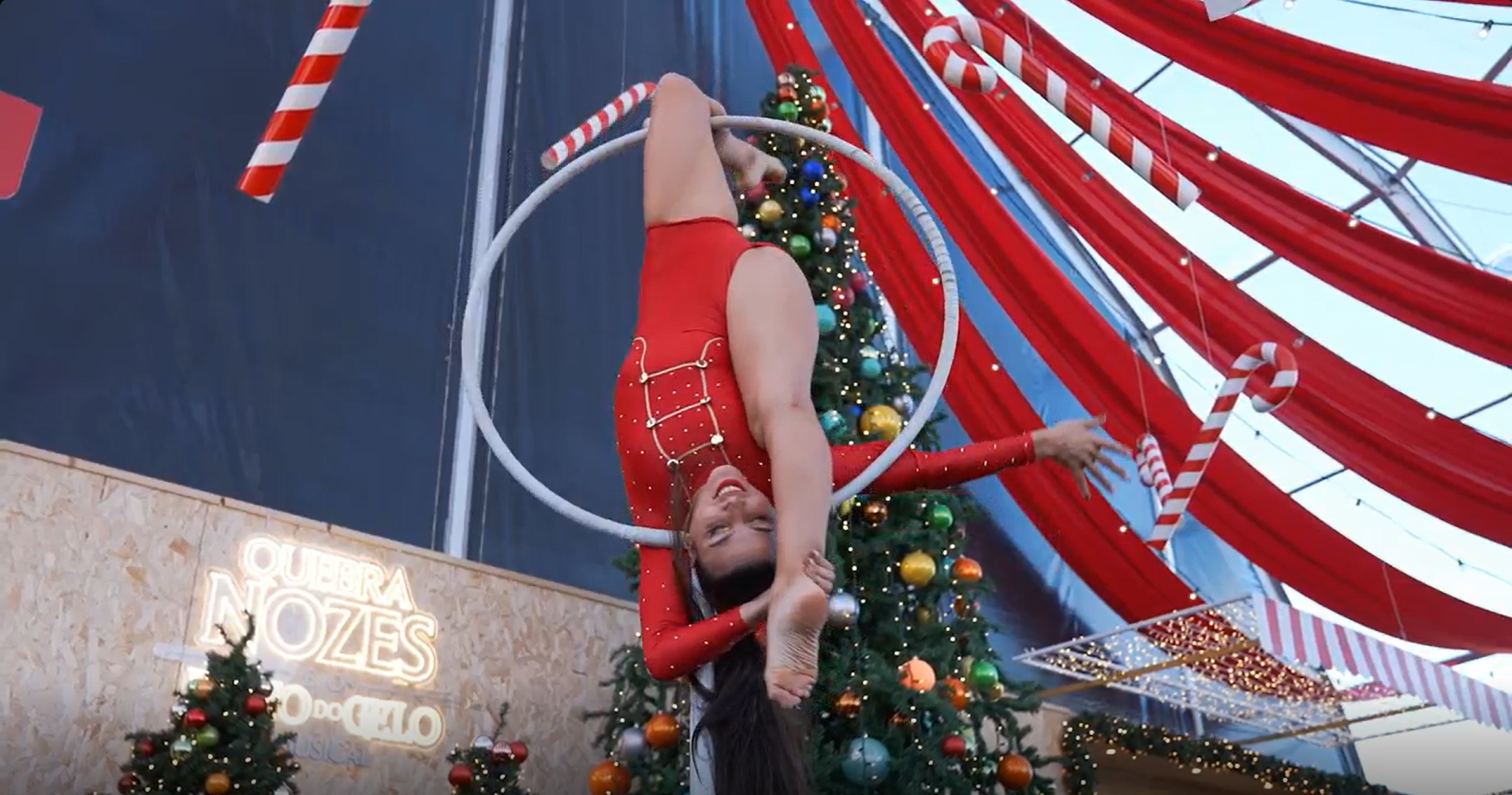 Mulher praticando acrobacias em um aros natalino decorado com árvores de Natal e enfeites, em ambiente de época natalina com luzes e decorações vermelhas e de doces tradicionais.