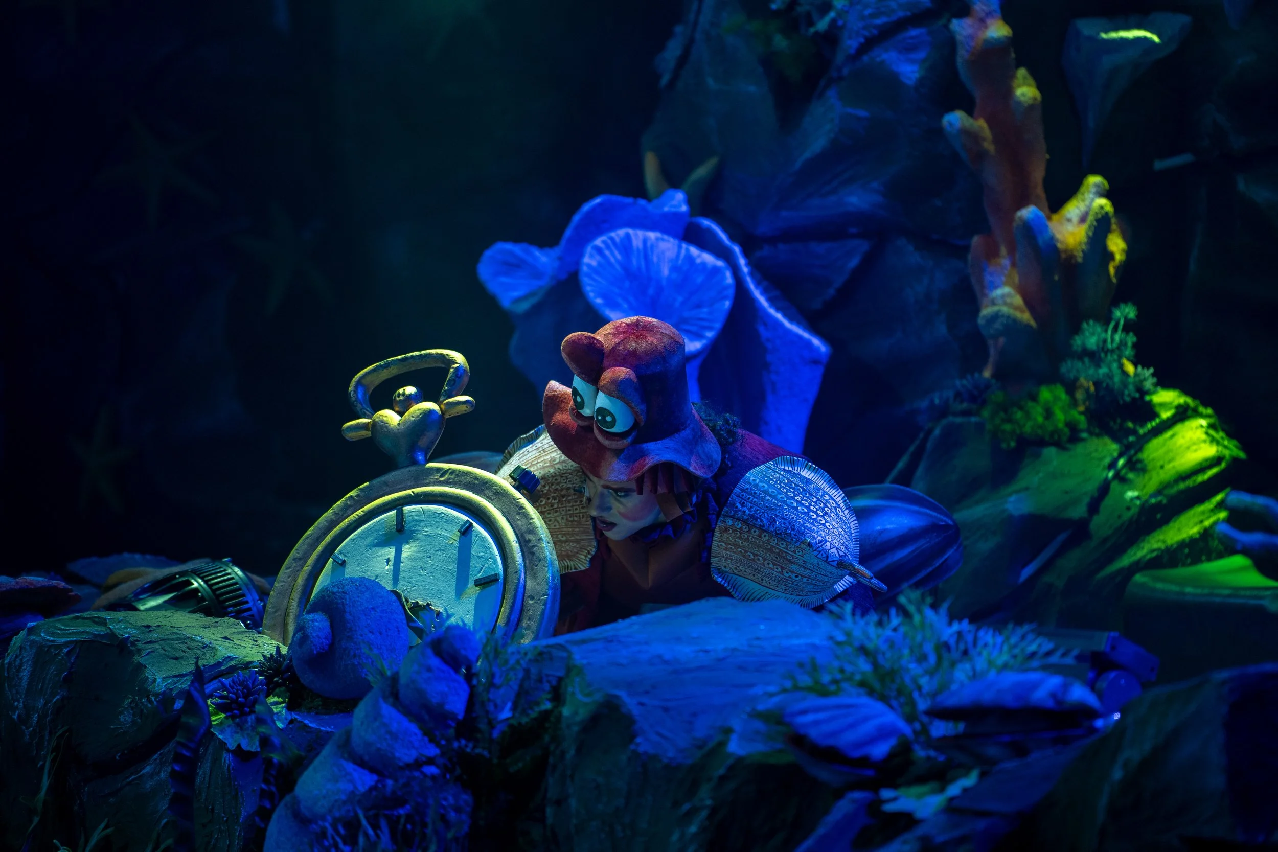 Cena de "A Pequena Sereia No Gelo" com Horácio Crustáceo junto a um relógio de bolso gigante, num cenário submarino iluminado em tons de azul. Corais, conchas e elementos marinhos envolvem a cena, criando uma atmosfera mágica inspirada no fundo do ma