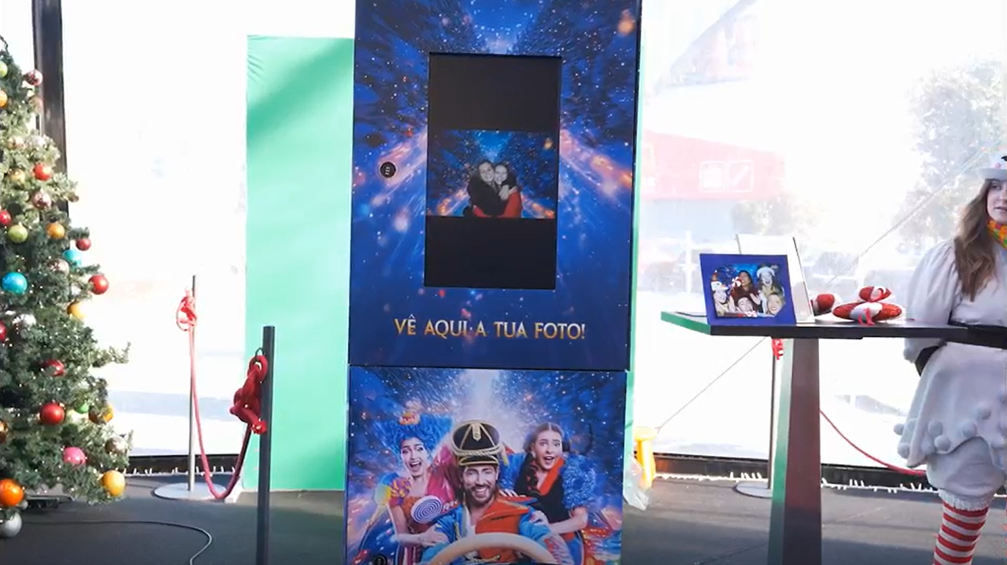 Estação de fotos natalina com painel azul, árvore de Natal decorada à esquerda, uma pessoa vestida de Papai Noel à direita, uma mesa com presentes embrulhados, uma tela de fotos, e uma decoração festiva ao fundo.