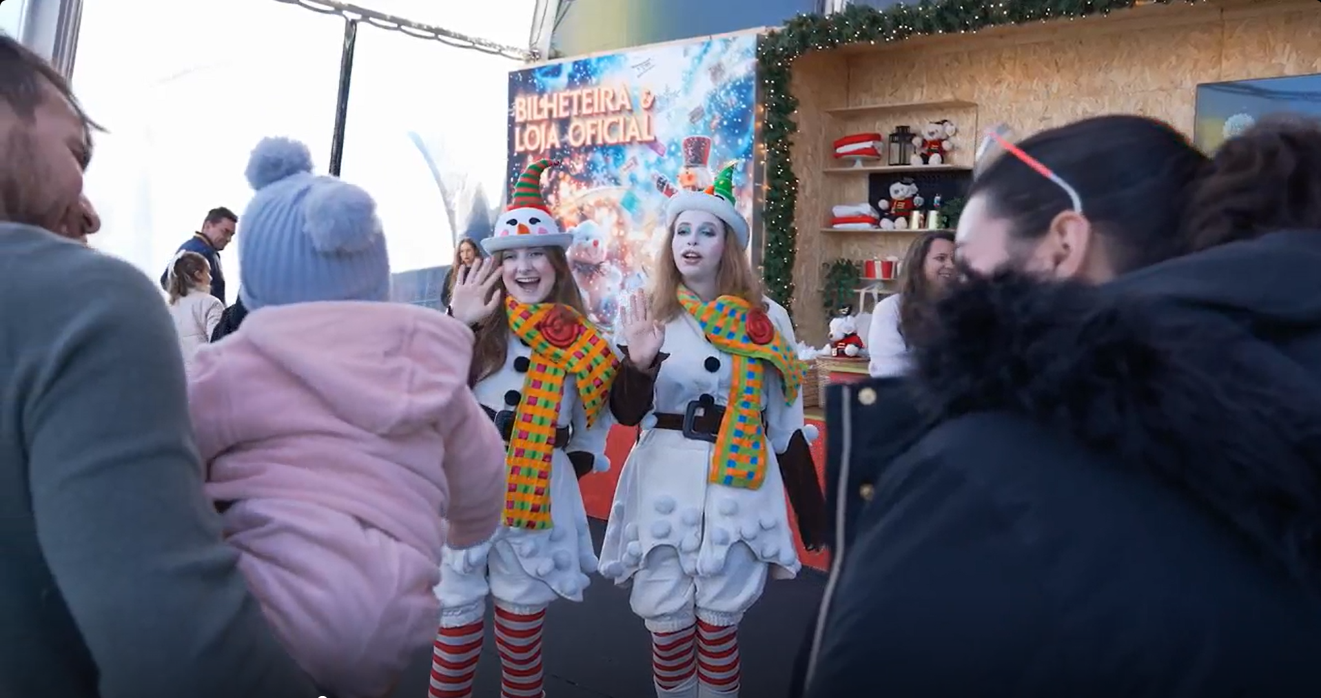 Duas pessoas vestidas de palhaço de neve com chapéus de Papai Noel e cachecóis coloridos, cumprimentando crianças e adultos em uma festa de Natal com decoração festiva ao fundo.