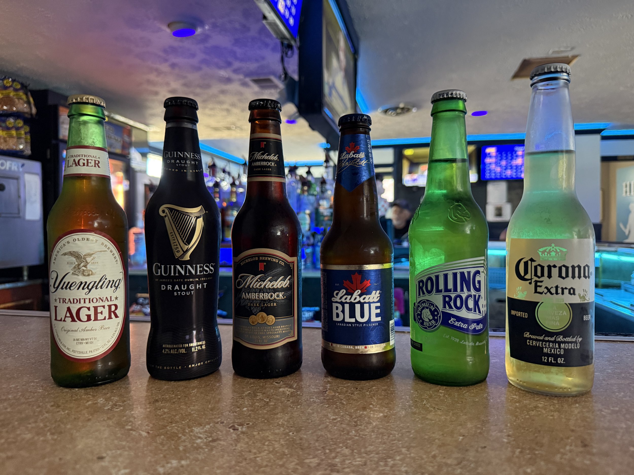 Various beer bottles lined up on a bar counter with a blurry background of a television and bar interior.