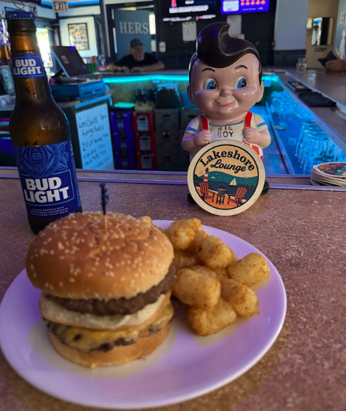 A plate with a cheeseburger and fried tater tots on a bar counter, a Bud Light beer bottle beside it, a smiling cartoon boy figurine holding a Lakeshore Lounge sign, in a bar or lounge setting.