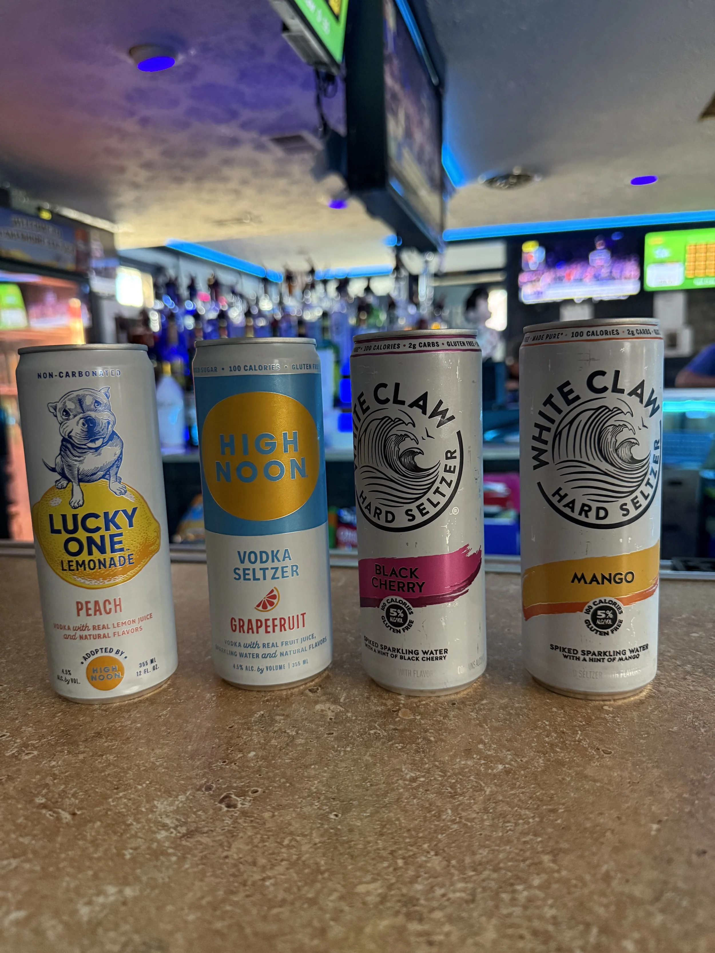 Four cans of flavored seltzer water on a bar counter, with a blurry bar scene in the background. The cans are labeled Lucky One Lemonade Peach, High Noon Grapefruit, White Claw Black Cherry, and White Claw Mango.