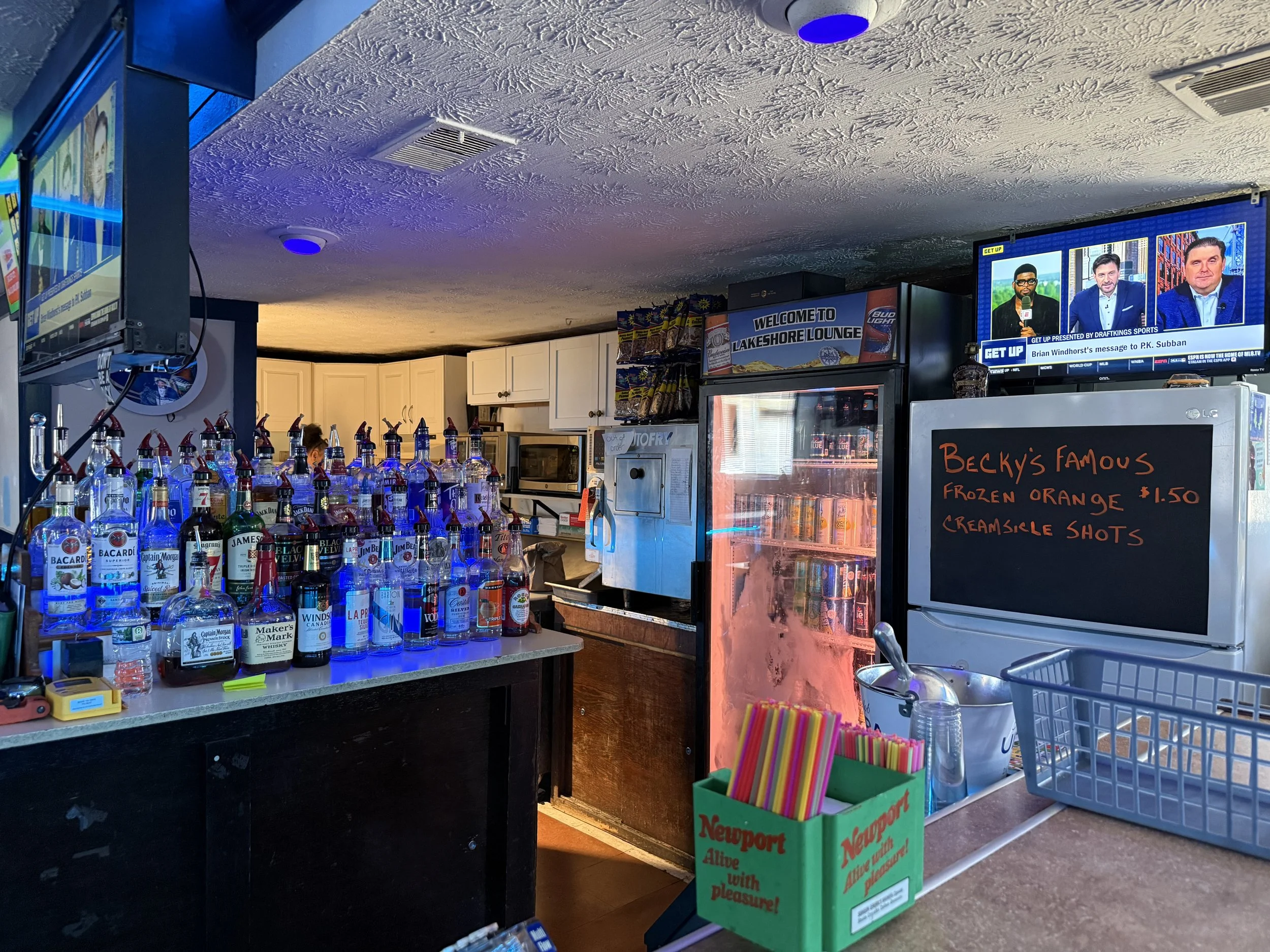 Bar inside a restaurant or bar with various alcohol bottles on the counter, a blackboard menu listing drinks, a TV showing a sports program, and a sign welcoming to Lakeshore Lounge.