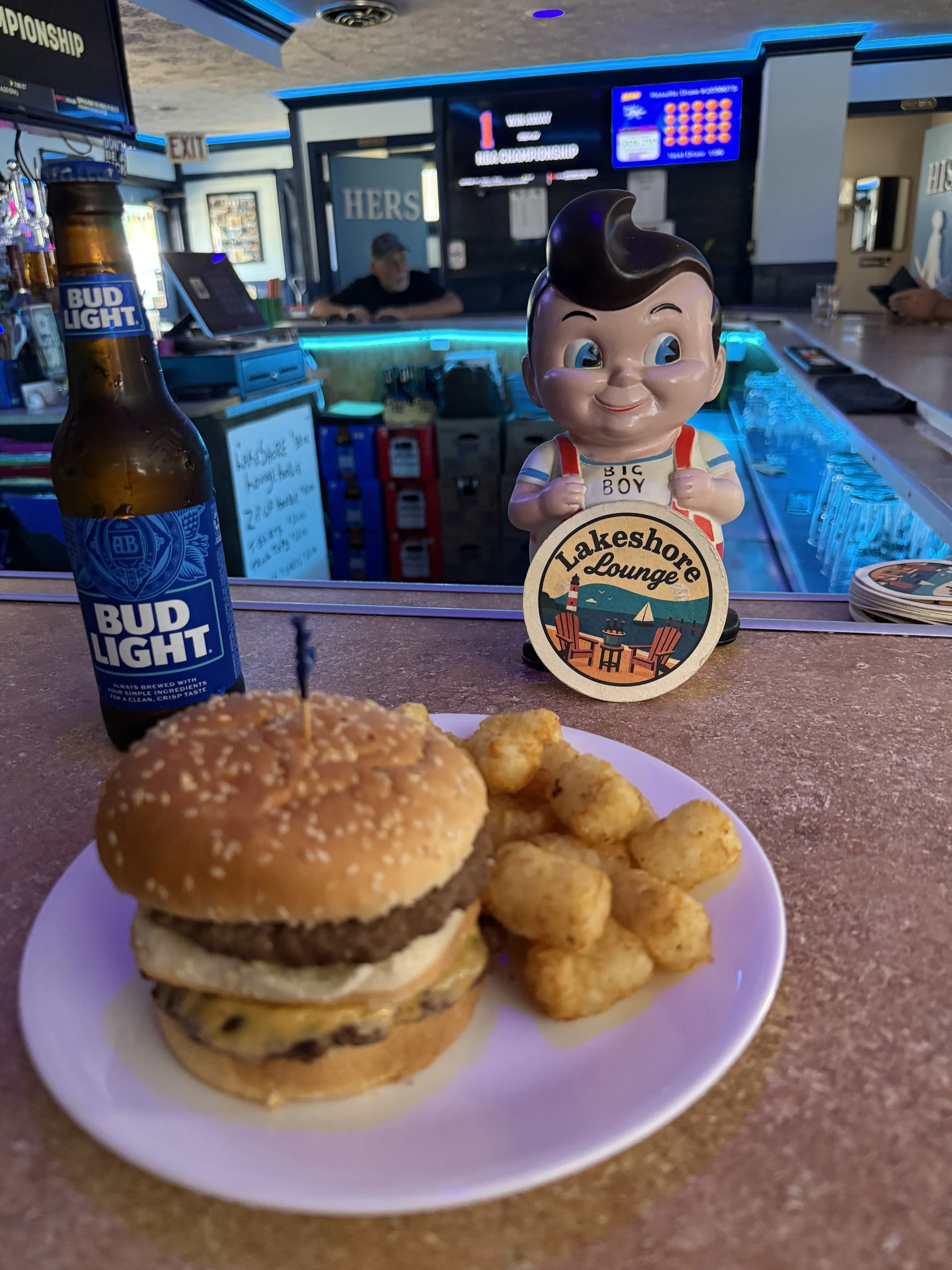 Close-up of a cheeseburger on a white plate with seasoned potato wedges on the side, in a restaurant setting with a Bud Light beer bottle, a statuette of a cheerful boy with a sign that says Lake Shore Lounge, and a bar in the background.