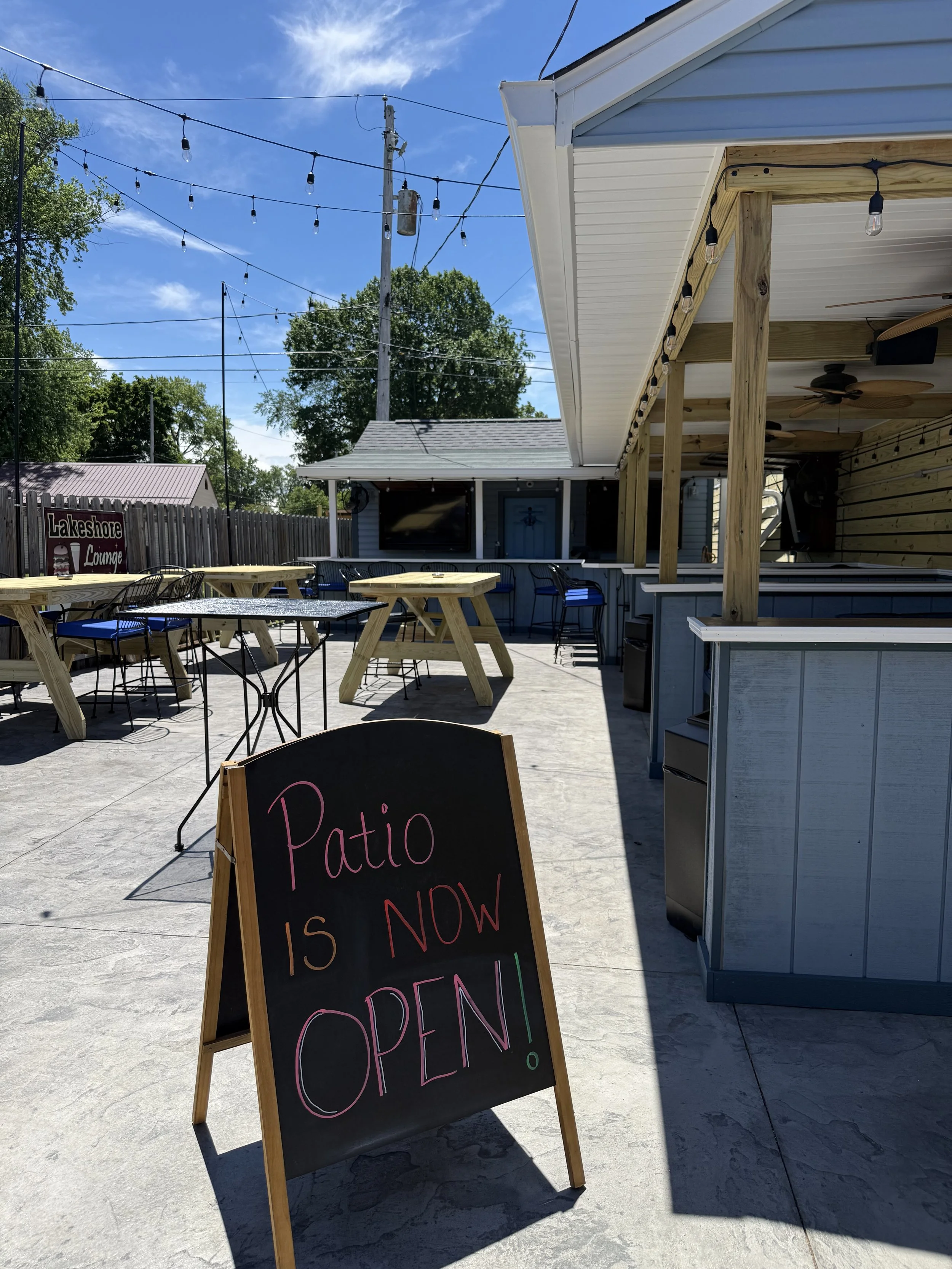 An outdoor patio with tables and chairs, string lights, a sign that reads 'Patio is now open,' and a building with a covered area under a blue sky.
