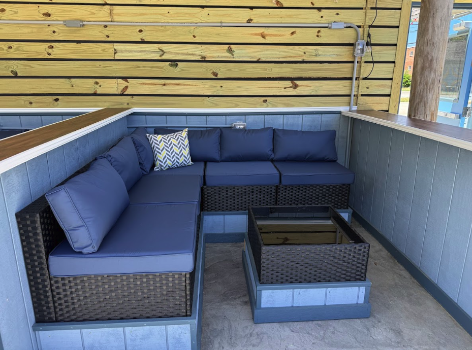 A corner outdoor seating area with a L-shaped dark wicker sofa with blue cushions and a decorative pillow, a matching coffee table with a glass top, set against a wooden wall with electrical wiring visible.