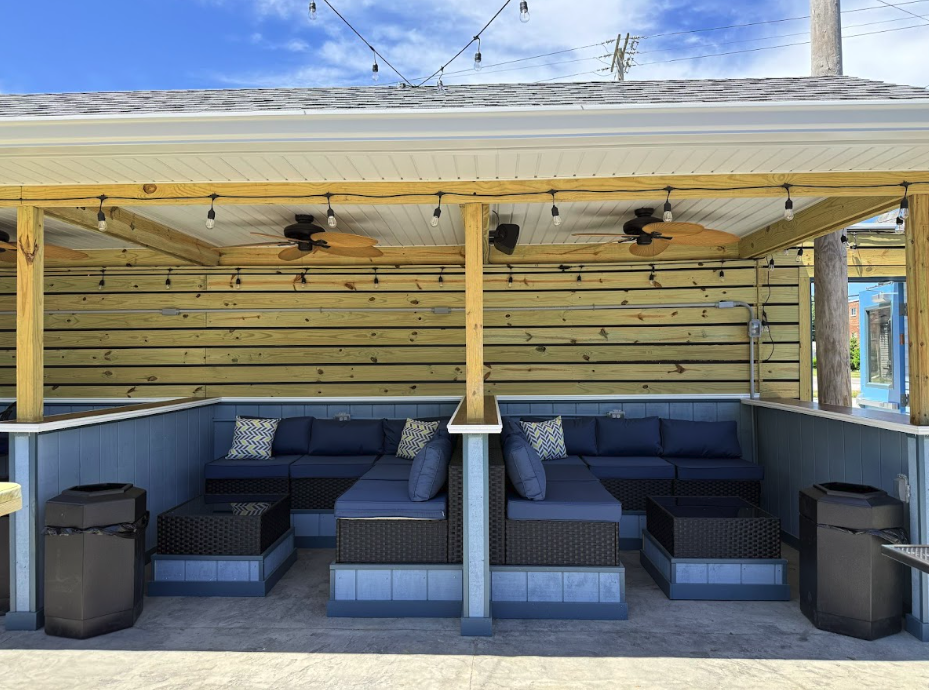 Outdoor seating area with blue cushions, pillows, and wicker furniture under a wooden roof with string lights, ceiling fans, and a wooden wall backdrop.