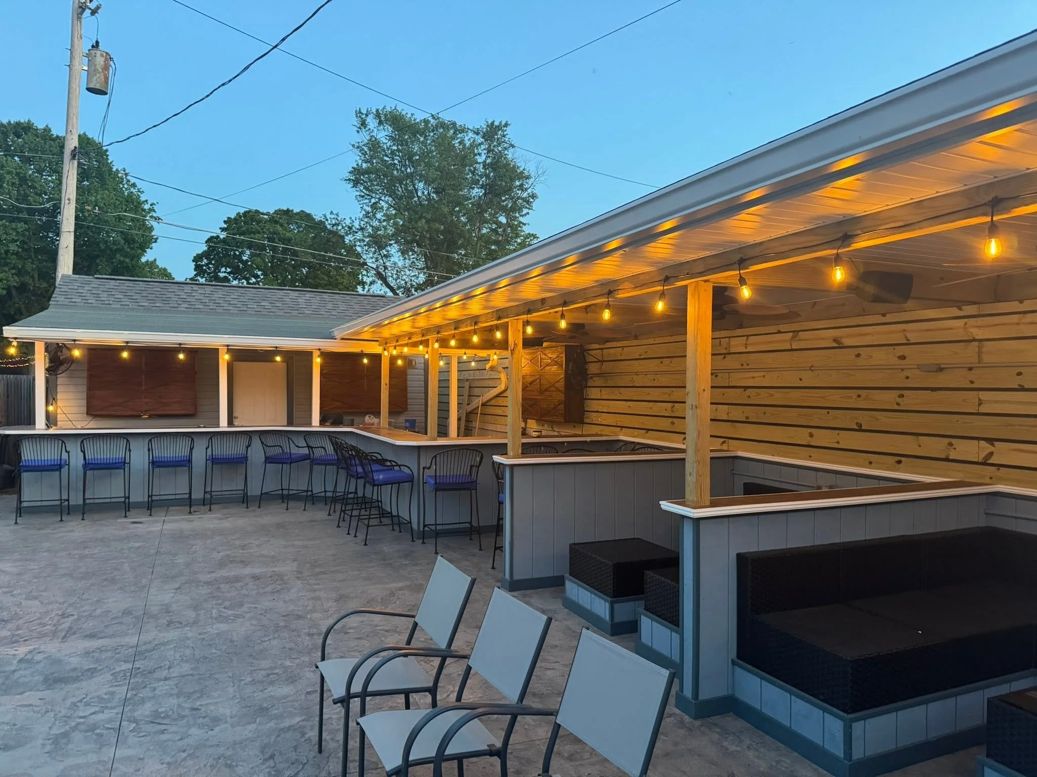 An outdoor patio area with barstools along a curved counter, string lights hanging from a wooden pergola, and a wooden privacy fence. There is a house with a sloped roof and trees in the background.