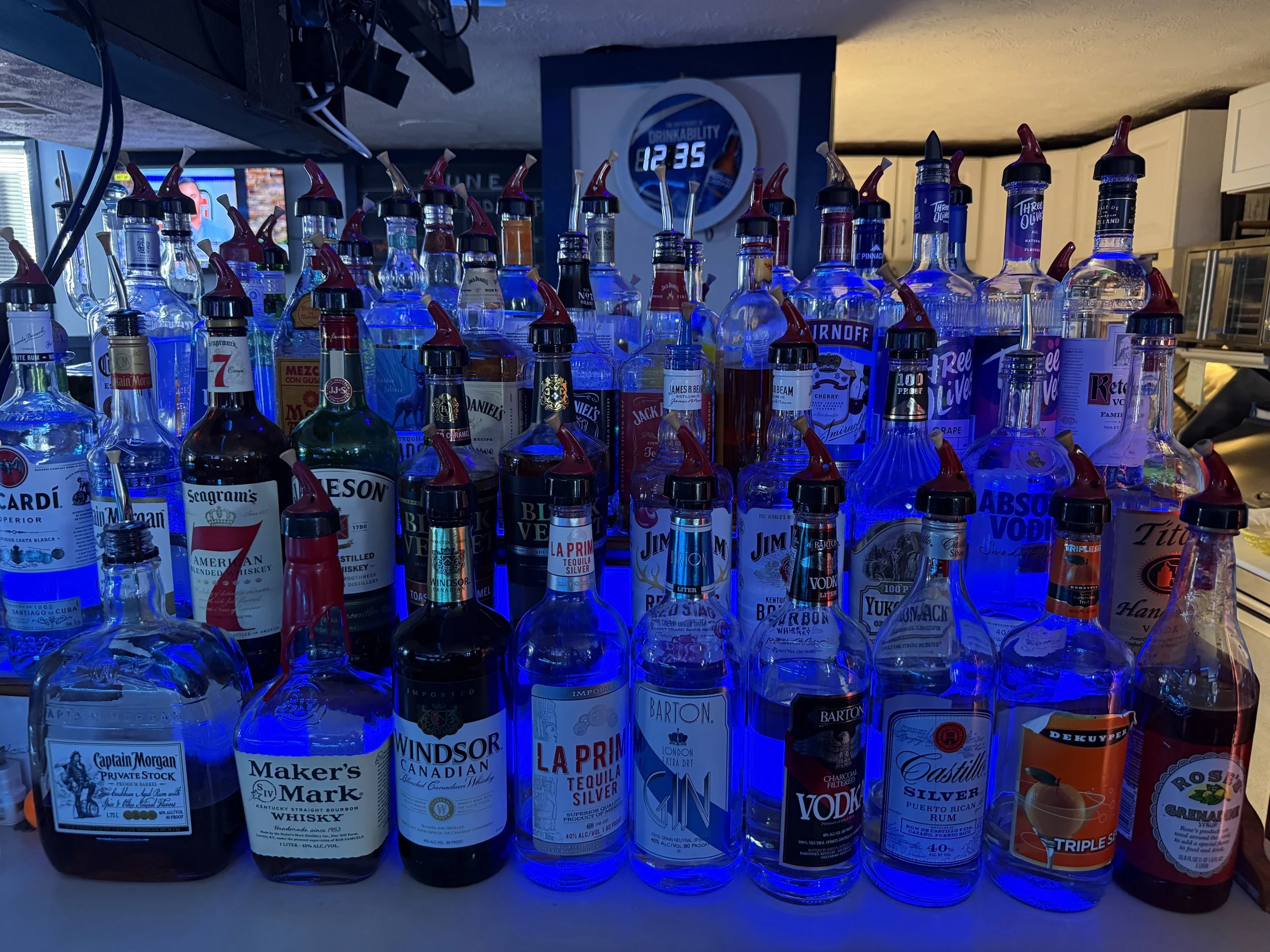 A collection of various liquor bottles, primarily vodka, displayed on a bar with a blue backlight. In the background, there is a wall clock showing the time as 12:35 and a sports channel on a television.