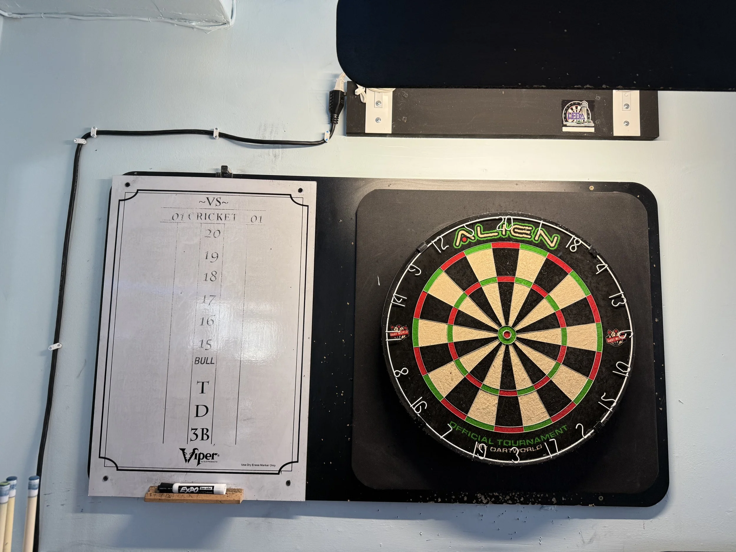 Dartboard with colorful segments on a black stand, a whiteboard with game scores, and three white markers on a light blue wall.