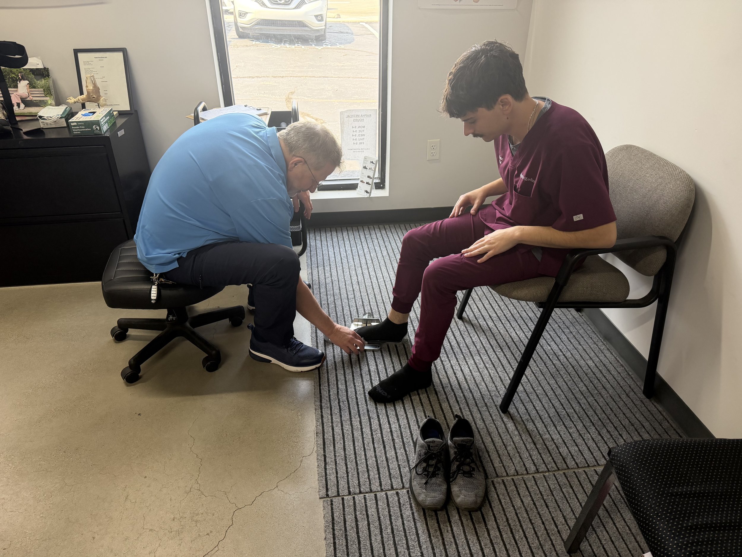 A doctor is sitting on a chair, bending forward to examine a young man seated nearby in a medical office. The young man's right foot is being examined, and he is wearing maroon scrubs with a name badge. The room has a window revealing a parking lot outside, and medical posters and supplies are visible on a shelf.