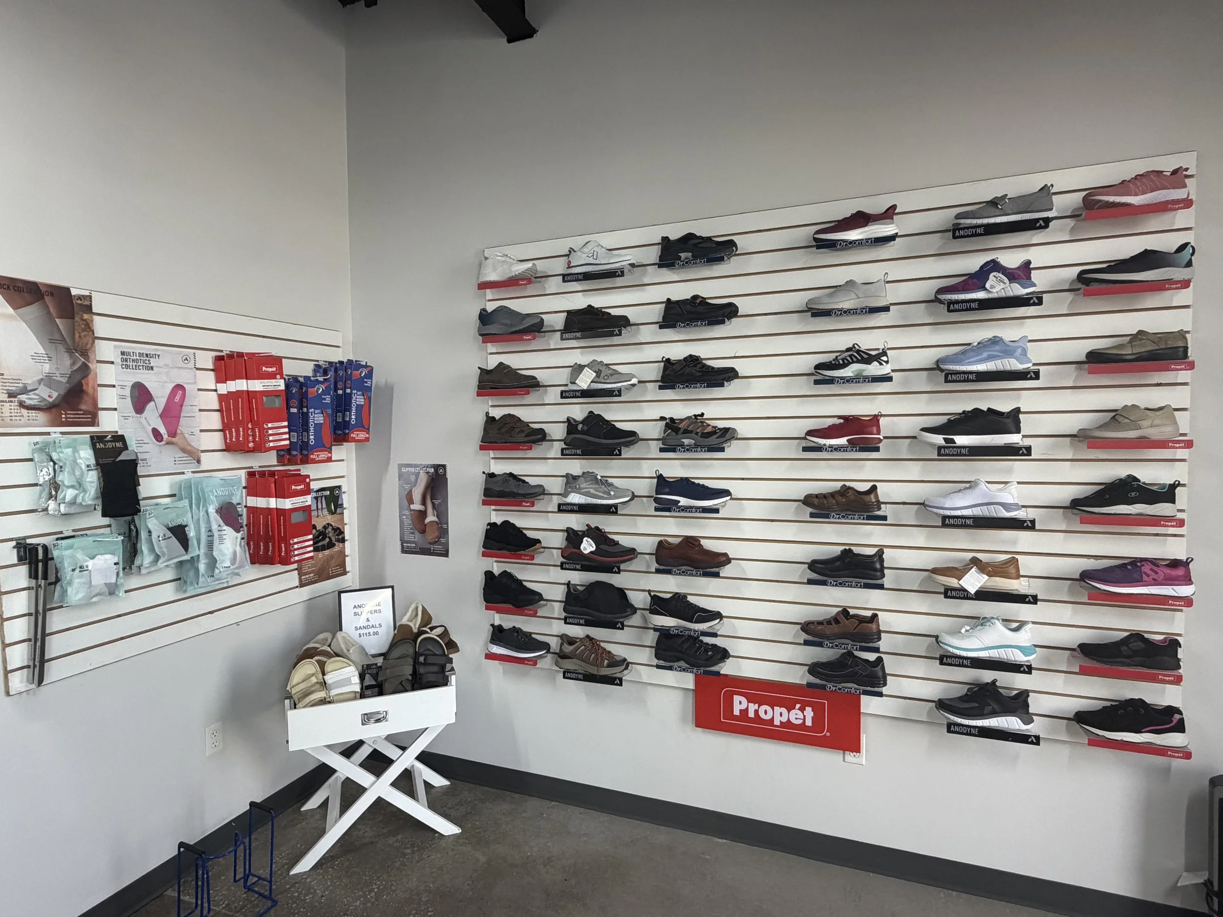 Display of athletic shoes on a wall in a store, with various brands and styles organized on horizontal slatwall shelves and some promotional posters and products visible nearby.