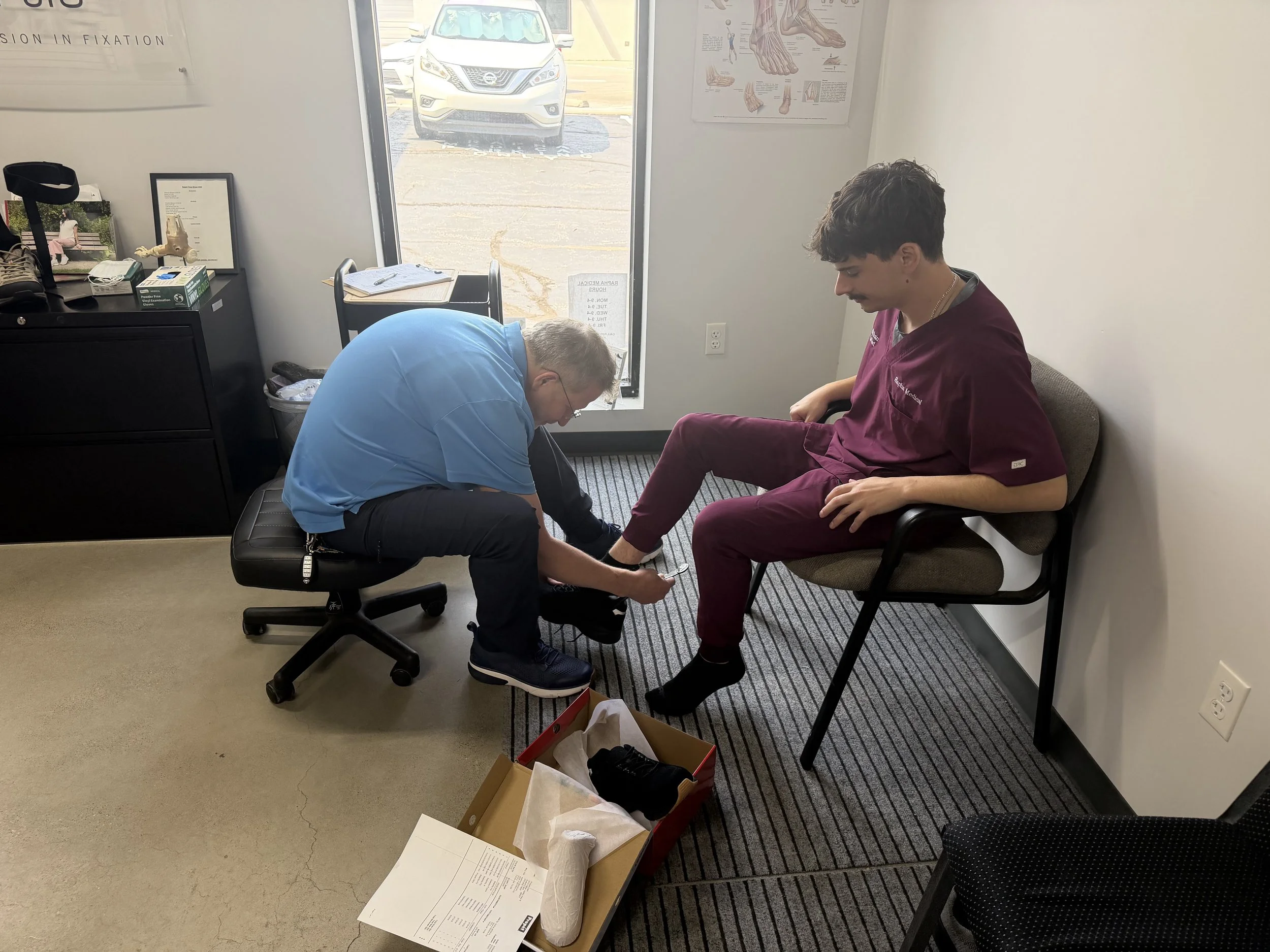 A young man in maroon medical scrubs sits in a waiting room, getting his shoe tied by an older man who is kneeling down. The older man is wearing a blue shirt and glasses. On the floor are a shoe box with black shoes, a crumpled paper bag, and some documents. Behind them is a window showing parked cars outside, and walls with medical posters and a sign.