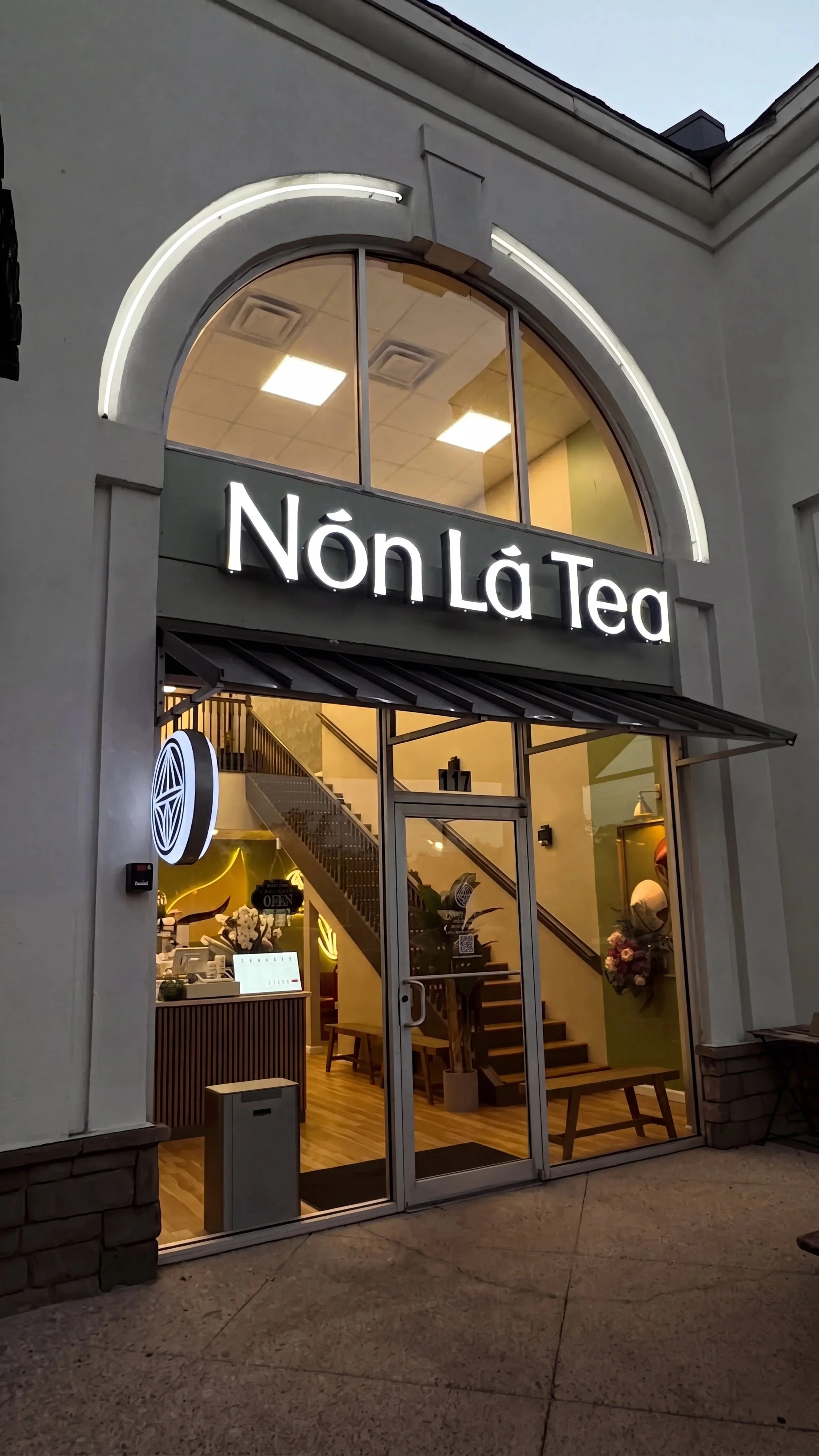 The storefront of a tea shop named Nòn Lá Tea with large glass windows, a neon sign, and interior visible including stairs, flowers, and seating.