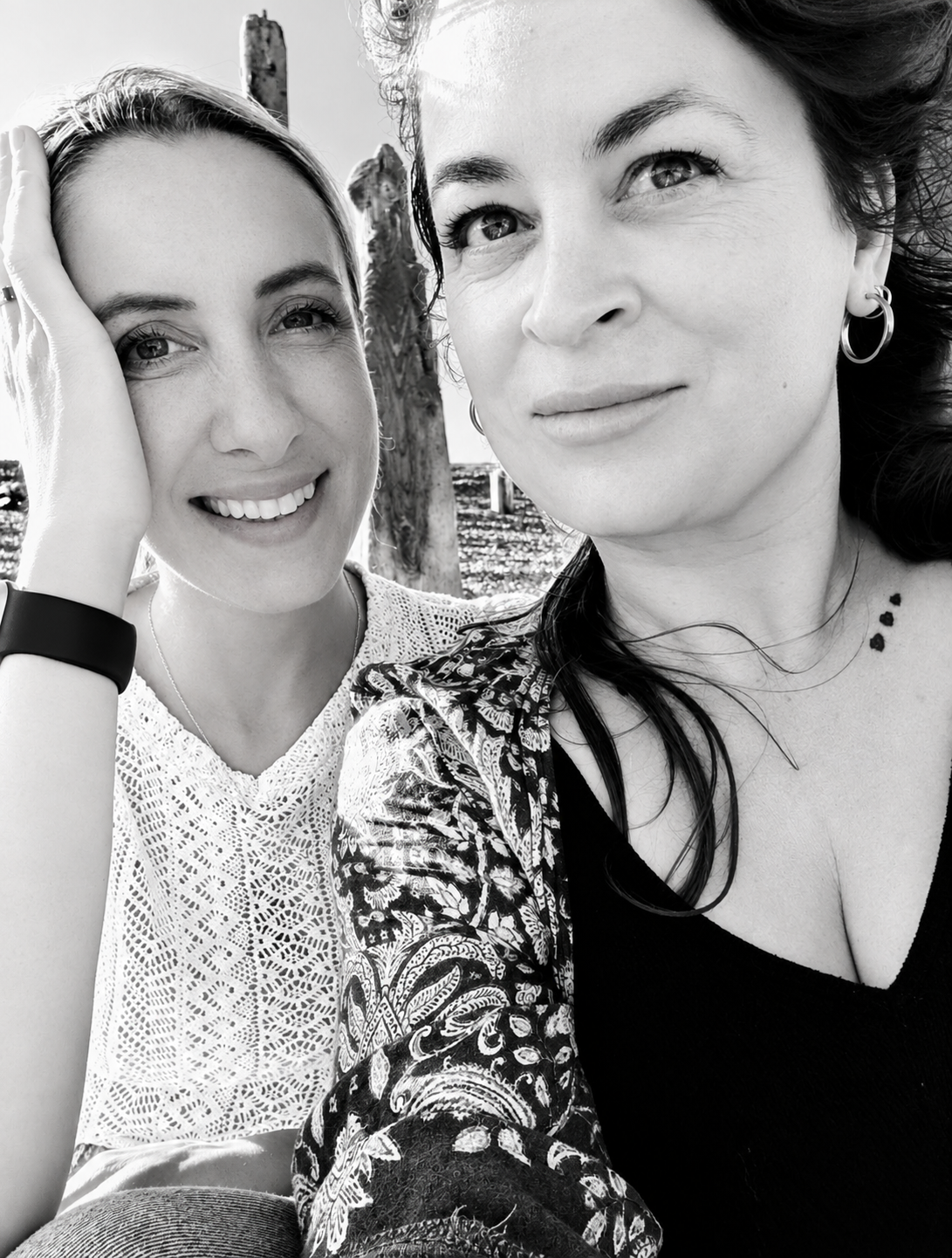 Two women smiling outdoors, close-up black and white photo, one with curly hair and earrings, the other with straight hair and a lace top.