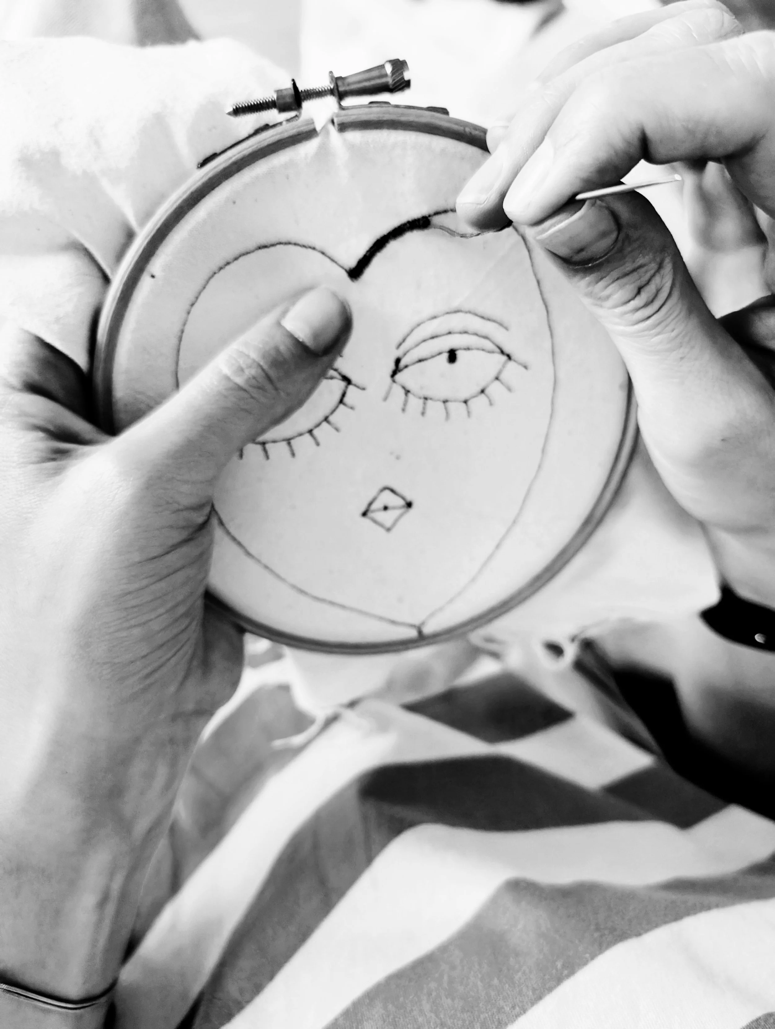 Close-up of hands holding an embroidery hoop with a drawing of a face, and a person using a needle to stitch black thread in the eye area.
