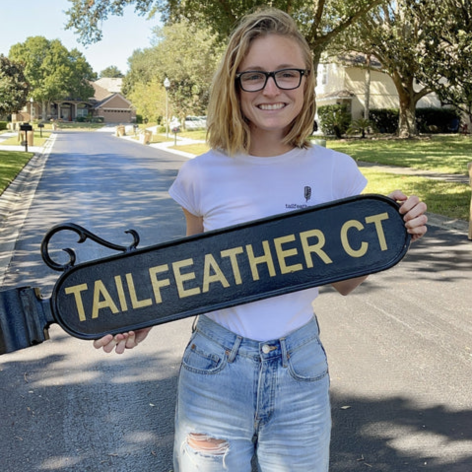 Woman with blonde hair and glasses standing on a suburban street, smiling and holding a street sign that says "TAILFEATHER CT."