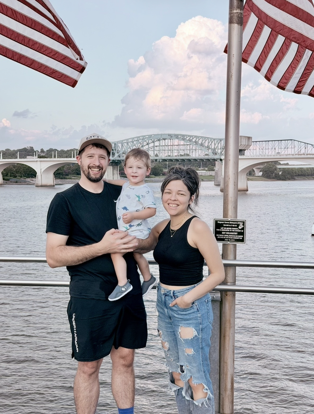 A smiling family of three standing on a riverside boardwalk with American flags overhead, a bridge in the background, and cloudy sky.