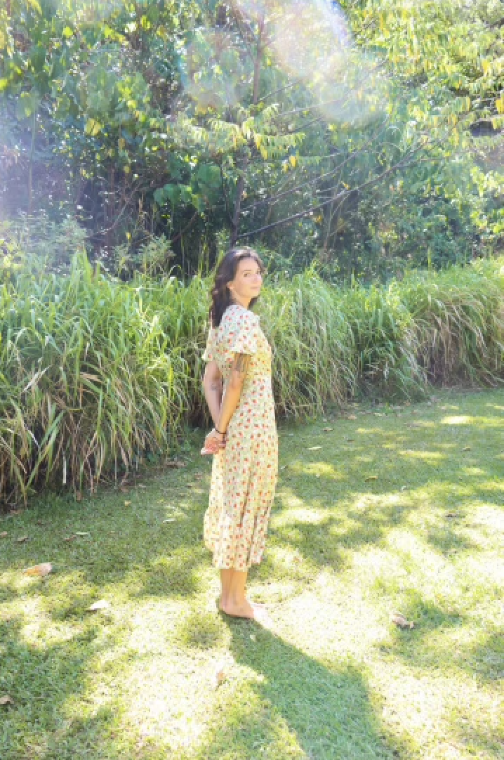 A woman in a floral dress standing outdoors on grass, with green bushes and trees in the background.