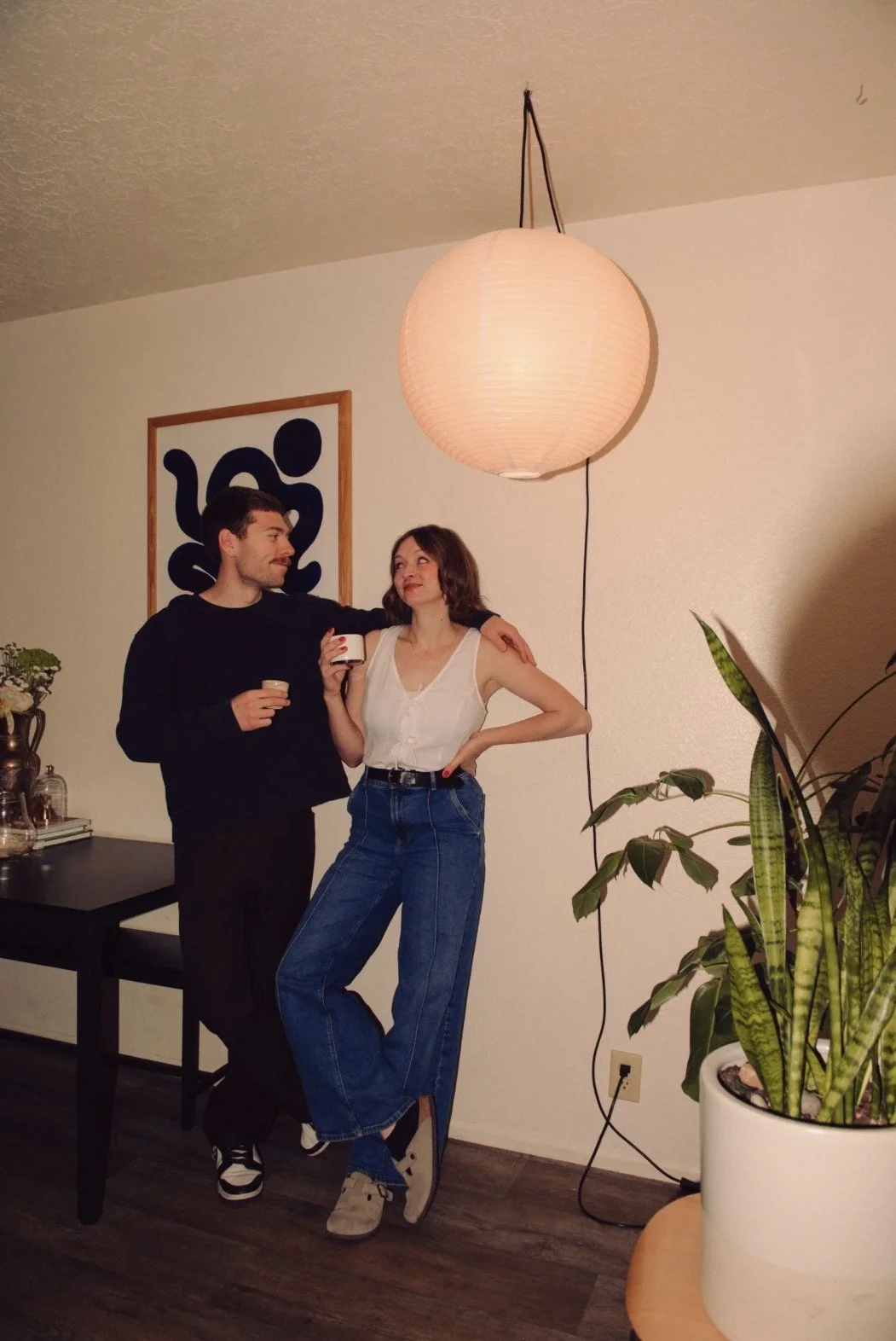 A man and woman stand together in a warmly lit room, holding coffee cups, with the man resting his arm on the woman's shoulder. They are near a table with decorative items, and there is a potted plant in the foreground. A large paper lantern hangs from the ceiling, and a framed abstract black design is on the wall behind them.