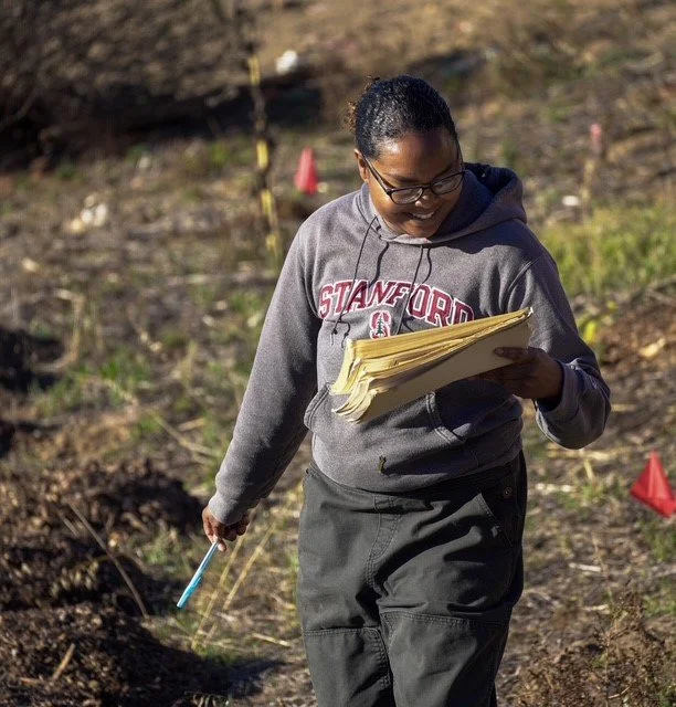 A woman wearing glasses and a gray Stanford hoodie, holding a clipboard with papers, walking through a field with red flags.