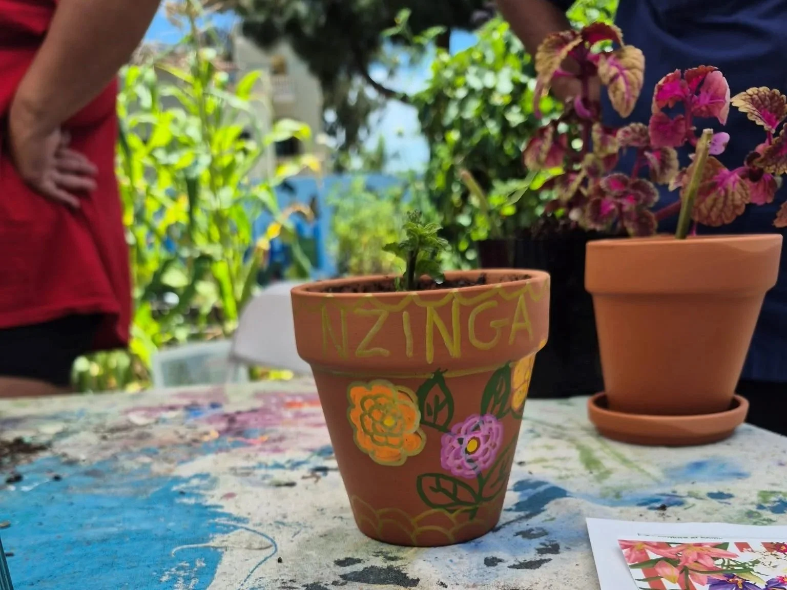 Colorfully decorated terracotta flower pot with the name 'NZINGA' and floral designs, placed on a table with other potted plants and people in the background.
