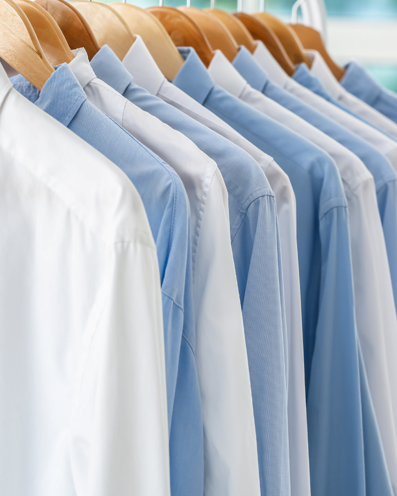 Multiple collared dress shirts in white and light blue hanging on wooden hangers in a row.