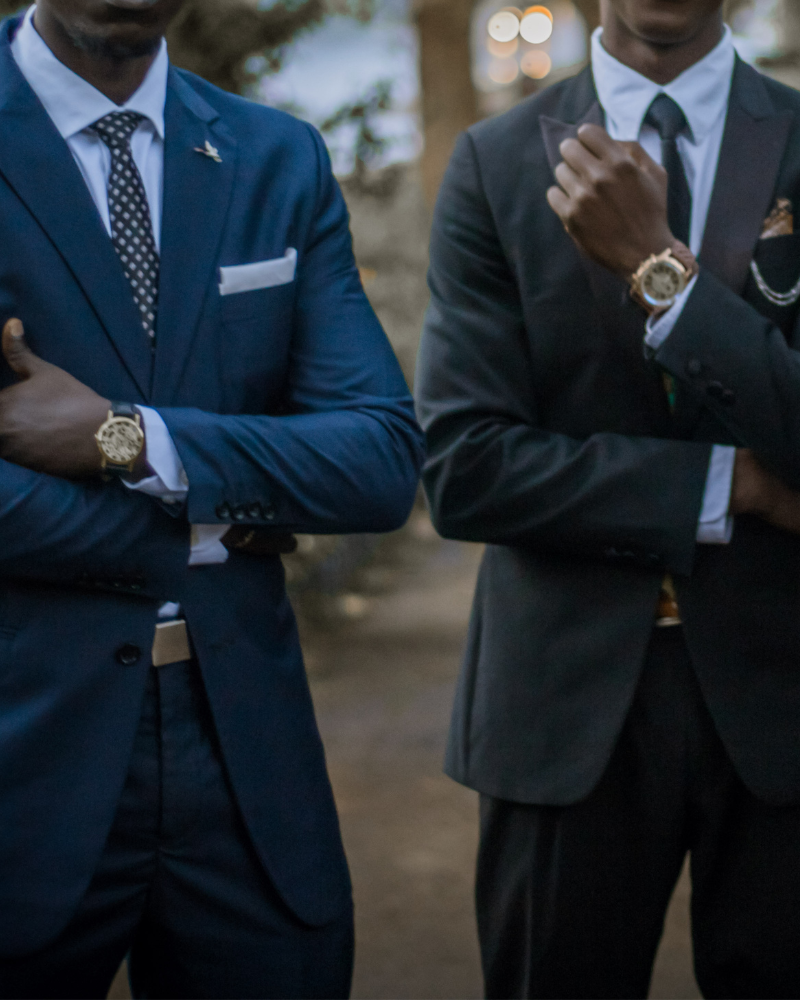 Two men dressed in formal suits with watches, standing outdoors with arms crossed and hand on chin, in a dimly lit setting.