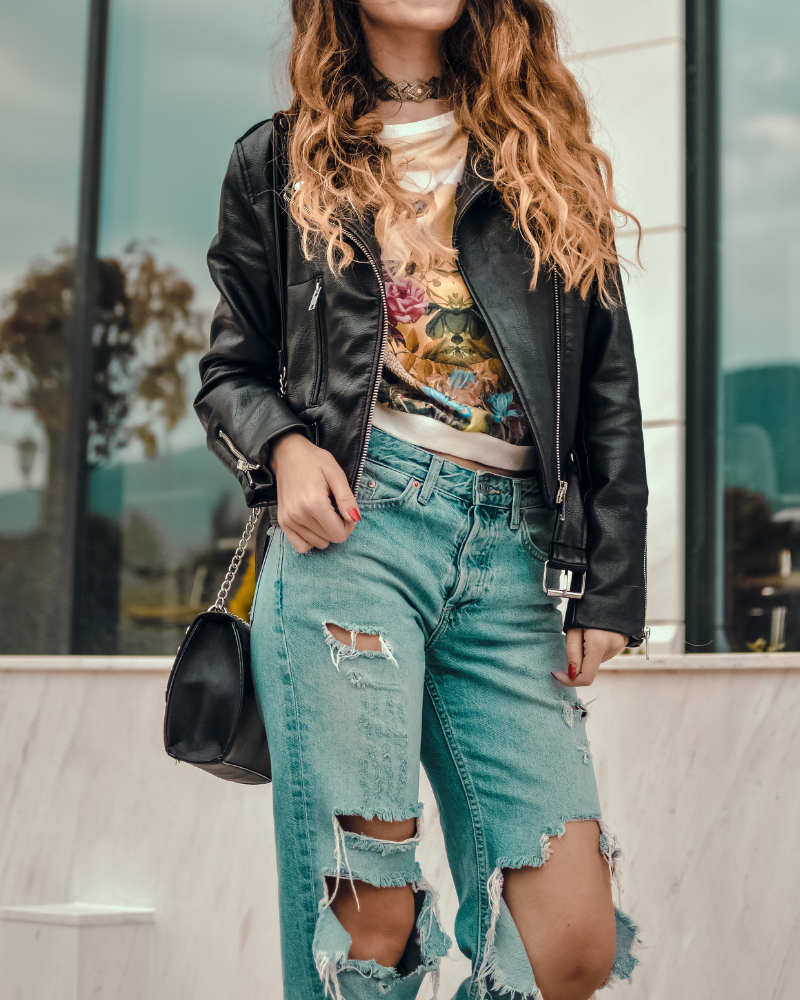 Person with long wavy hair wearing a black leather jacket, floral shirt, ripped jeans, and holding a black purse standing outdoors.