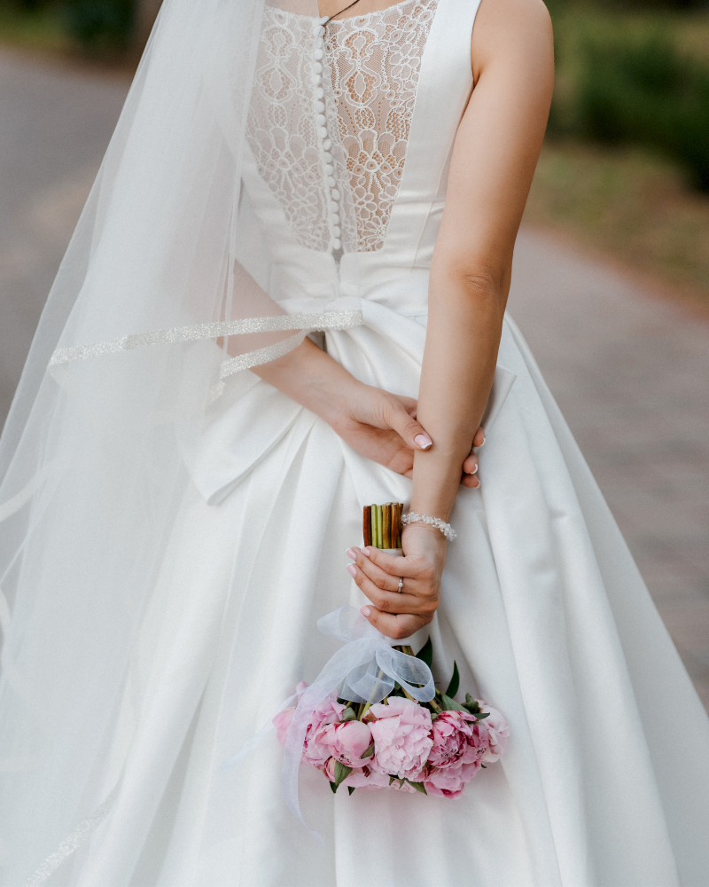 A bride in a white wedding dress holding a bouquet of pink peonies, standing outdoors.