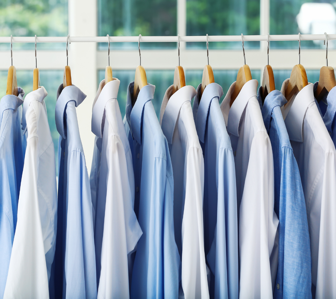 A row of men's dress shirts in shades of blue and white hanging on wooden hangers on a metal clothing rod, with a window in the background letting in natural light.