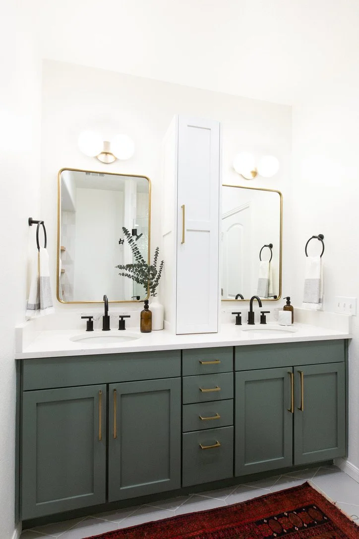 Modern bathroom vanity with two mirrors, black faucets, a white countertop, green cabinets with gold handles, and two wall mounted light fixtures.
